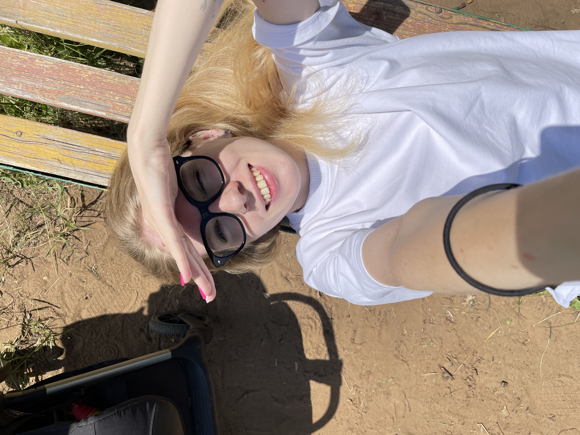 A young woman with blonde hair and sunglasses is lying on a wooden bench outdoors, holding her hand to her forehead to shield her eyes from the sun. She is wearing a white t-shirt and a black band around her forearm. A stroller is visible in the background. The ground is made of dirt and there is some grass. The image appears to have been taken on a sunny day. The specific location is unknown, but the scene suggests a park or outdoor recreational area.
