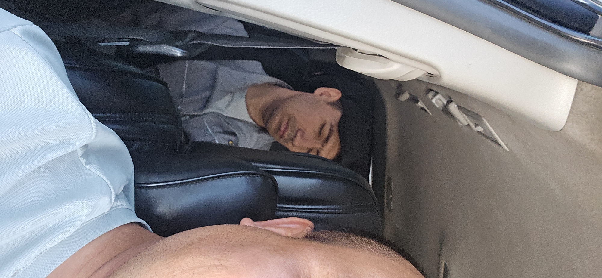 In the city of Guzar, Uzbekistan, a man wearing a grey work uniform and a black baseball cap is asleep in the back seat of a vehicle. The man is slumped against a dark-colored headrest, his eyes closed and his mouth slightly open. The interior of the car is visible, including a white door panel on the left and black leather seats. In the foreground, the shoulder and neck of another person, likely the photographer, are visible, wearing a light blue shirt. The scene appears to be a daytime interior shot.