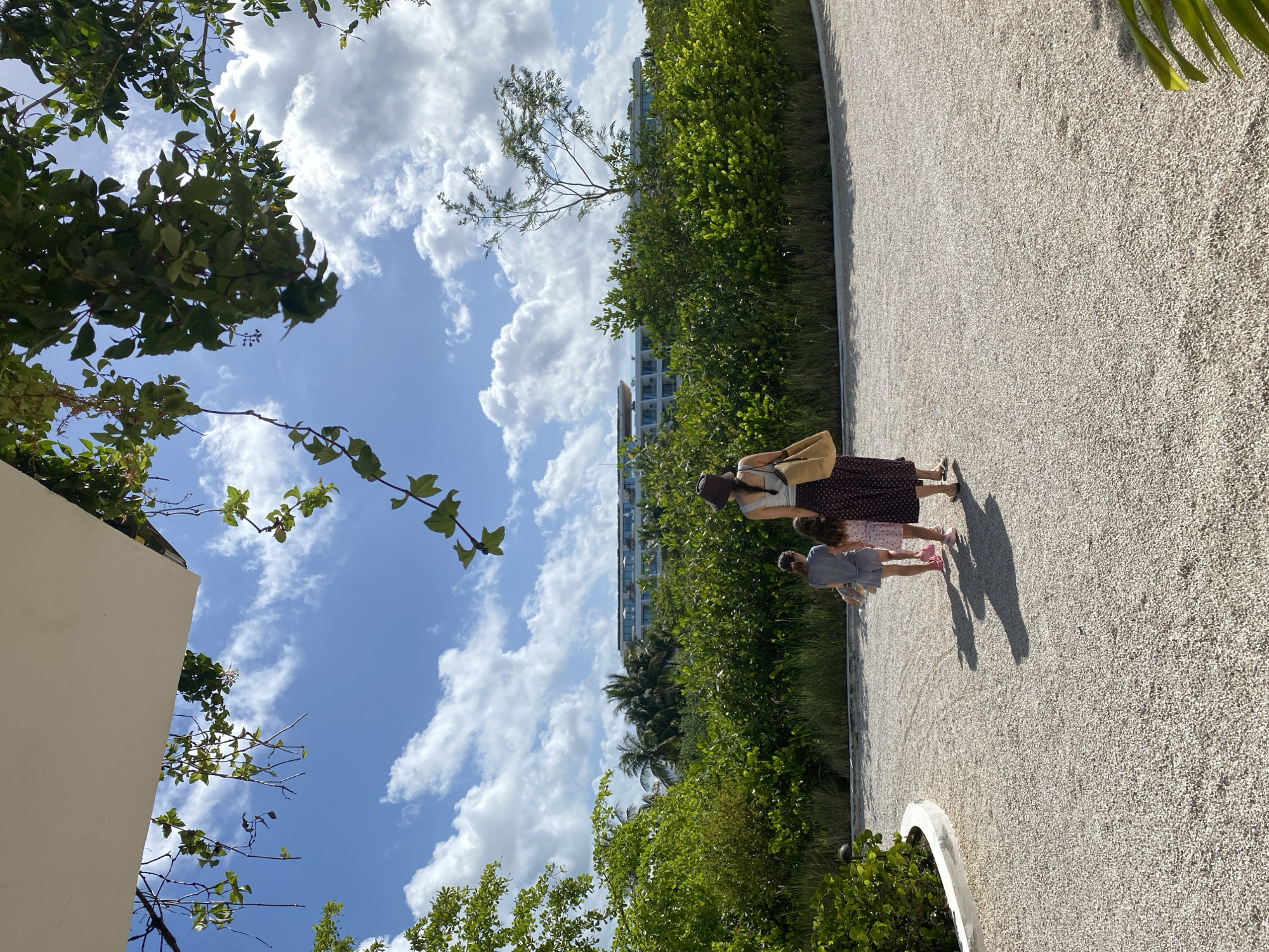 The image depicts an outdoor scene with a woman and two children walking along a gravel path in Marina Maroma, Mexico. Lush green vegetation lines the path, and a multi-story building is visible in the background amidst palm trees. The sky is blue with fluffy white clouds. The perspective is slightly angled upwards, capturing trees and foliage framing the sky.