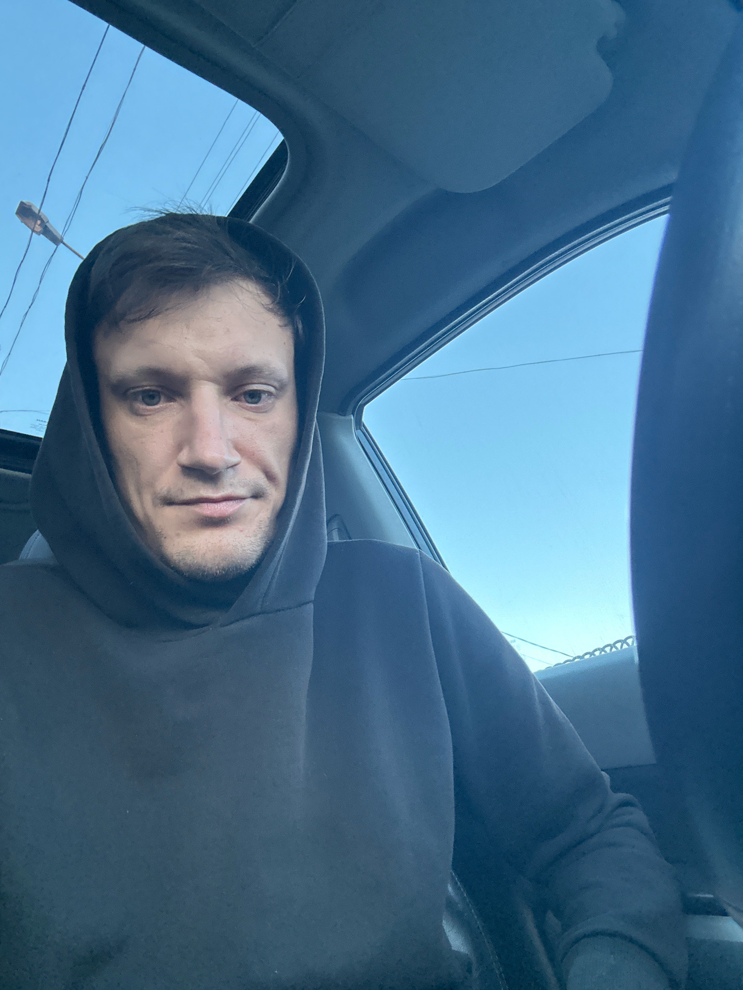 A man wearing a dark hooded sweatshirt is pictured inside a car, with the sunroof open and the blue sky visible above. The man's face is turned towards the camera, and he appears to be looking directly at it. The interior of the car is dimly lit, with the exception of the light coming through the sunroof. The background shows a bright blue sky with some utility wires and a street lamp visible. The image seems to be a selfie taken from the driver's seat of the car. There is no discernible information about the specific location within the city of Makiv, Ukraine.