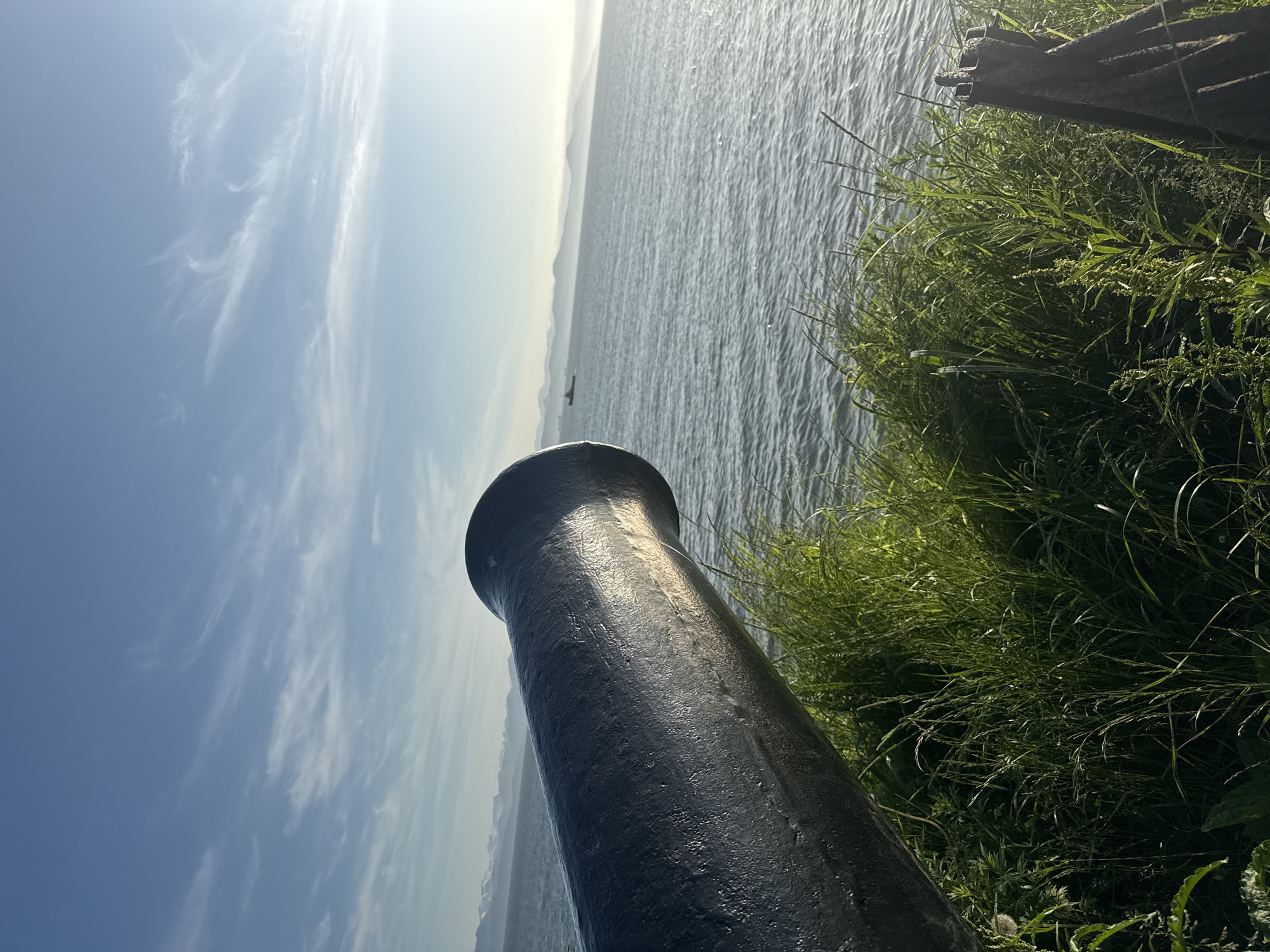 The image shows a close-up of the muzzle of a dark, weathered cannon pointing towards the sea. The cannon sits on a grassy embankment overlooking a calm, blue body of water. In the distance, a hazy horizon is visible with distant mountains or landforms. A small boat can be seen on the water. The sky is clear blue with wispy white clouds, and the sun is shining, casting a warm light. The scene appears to be an outdoor historical site or coastal defense location. The location is suggested to be Petropavlovsk-Kamchatsky, Russia, which is a port city on the Pacific coast known for its natural beauty and historical significance.