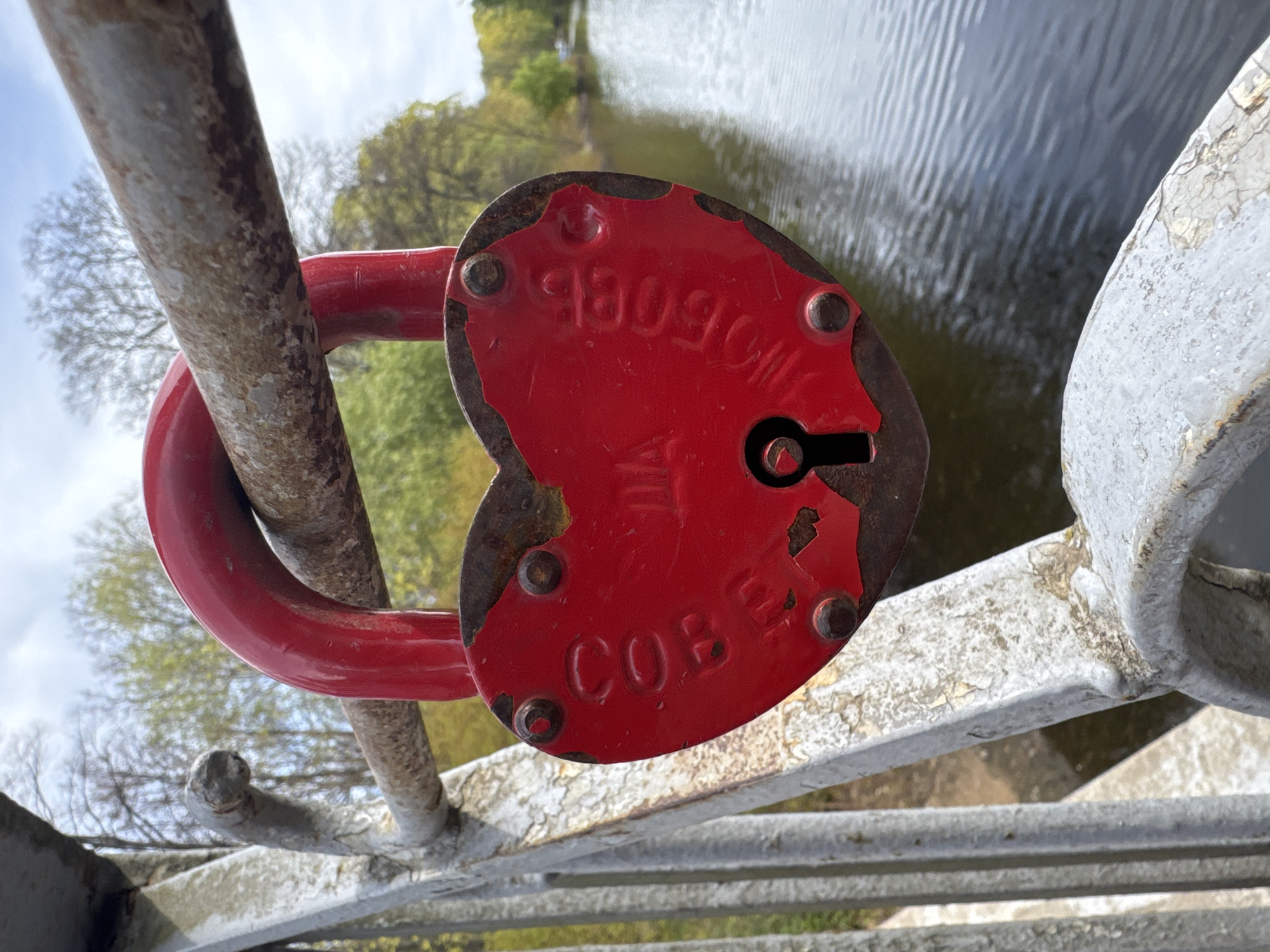 A close-up, slightly off-center, landscape-oriented photograph shows a red, heart-shaped padlock attached to a white metal railing over a body of water, likely a canal, in Saint Petersburg, Russia. The padlock has rusted edges and visible engraved Russian Cyrillic text, "СОВЕ" on the bottom and "93090M" on the top. The padlock shackle is secured around a rusty brown metal bar of the railing. The white metal railing has several horizontal bars and a decorative rounded element. In the background, a body of water reflects the sky and the nearby trees. The sky appears partially cloudy.