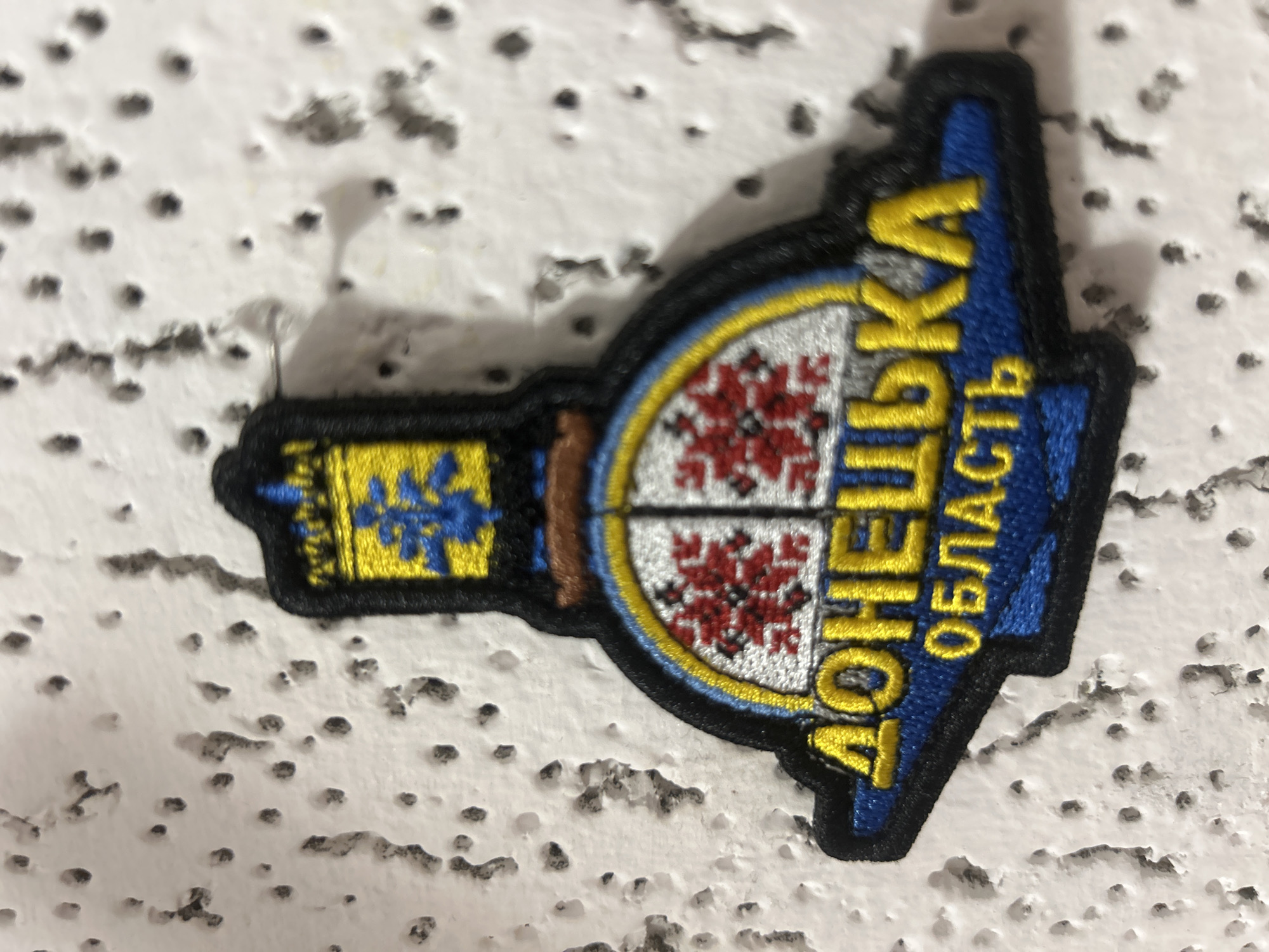 A close-up, eye-level, slightly blurred photograph captures a round-edged, black-bordered embroidered patch affixed to a textured white surface with small, dark divots. The patch depicts a stylized emblem in yellow, blue, and red. At the top, a yellow structure resembling a tower or monument with blue decorative elements is visible. Below this, a semi-circular section features a pattern of red embroidered designs against a white background, divided by blue lines. The bottom portion of the patch contains text in yellow embroidered letters against a blue background, reading "ДОНЕЦЬКА ОБЛАСТЬ". The patch is pinned to the wall with a small metal pin visible on the left side. The white surface appears to be a wall or textured material, common in interior or exterior urban settings. The lighting suggests an indoor environment, likely with artificial light. The image does not provide any information to determine the location within Krakow, Poland.