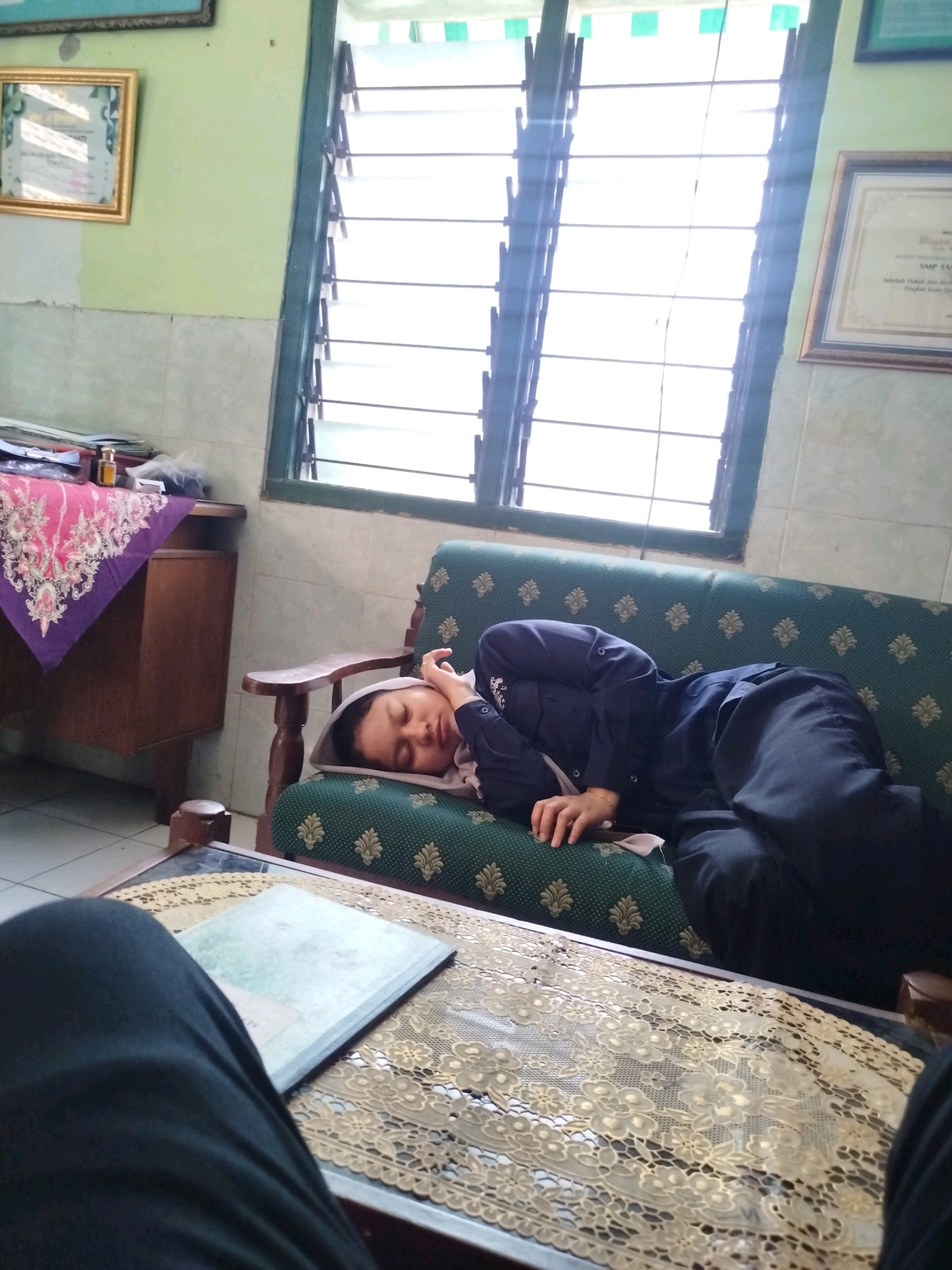 The image shows a person sleeping on a green couch in what appears to be a room. The person is wearing a dark blue uniform and is covered with a light-colored cloth. A wooden table with a decorative cloth and a book is in the foreground. The background includes a window with louvers, a wooden cabinet with a patterned cloth on top, and framed pictures on the wall. The environment suggests an indoor setting, possibly a school or office, given the uniform and framed certificates. Based on the visual cues, the scene is likely located in Madiun, Indonesia.