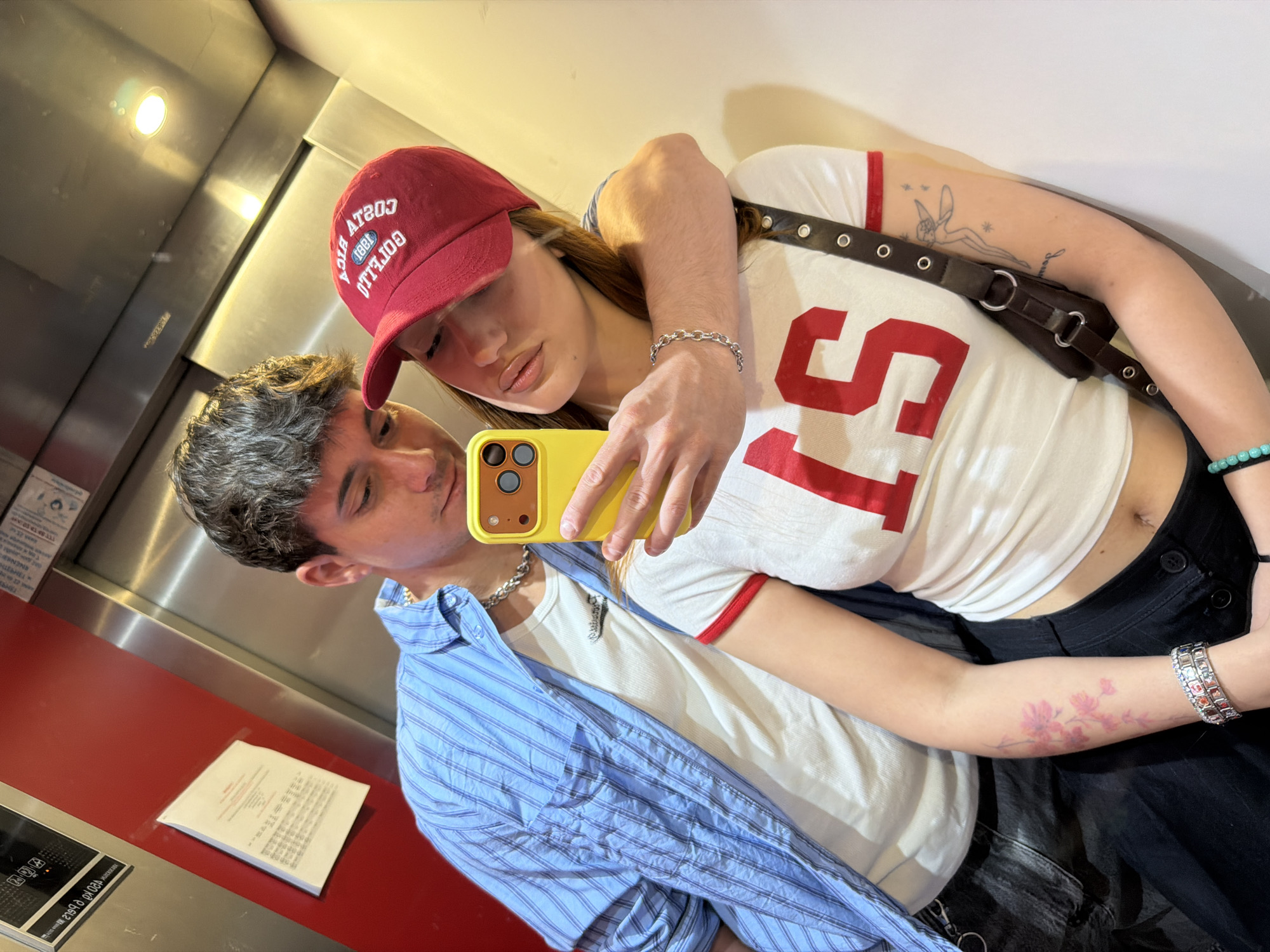 Two people are taking a selfie in what appears to be an elevator. The elevator has stainless steel walls and a red panel on one side. A document is visible on the red panel. The woman wears a red baseball cap with white text, a white shirt with red trim and a large red number "15", and black pants. She has a black purse slung over her shoulder, and tattoos are visible on her left arm. She holds a yellow smartphone. The man wears a blue striped shirt over a white undershirt. Both are wearing silver necklaces and bracelets. The location appears to be indoors, but the available information does not allow definitive placement in Sofia, Bulgaria.