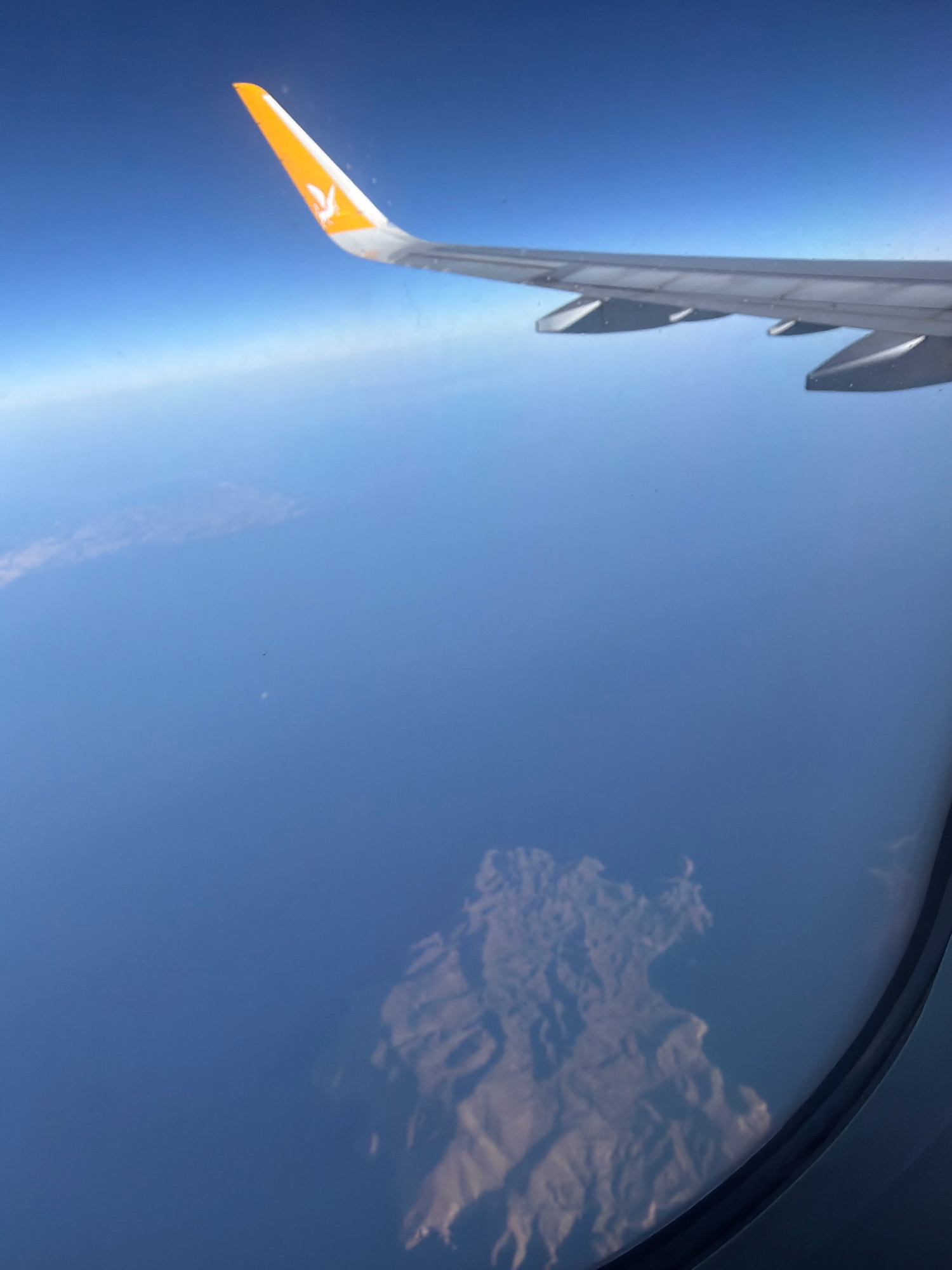 An aerial view from an airplane window shows a wing with a yellow and white logo against a clear blue sky. Below, a vast expanse of calm, deep blue water dominates the scene. In the distance, a faint, hazy landmass is visible. Closer to the foreground, a rugged island with visible mountainous terrain rises from the sea. The island appears arid with sharp, uneven contours, suggesting a geological landscape shaped by erosion. The overall environment is natural and remote, with no signs of human settlement or activity on the island. The image captures a moment of flight over a serene, undeveloped coastal region.