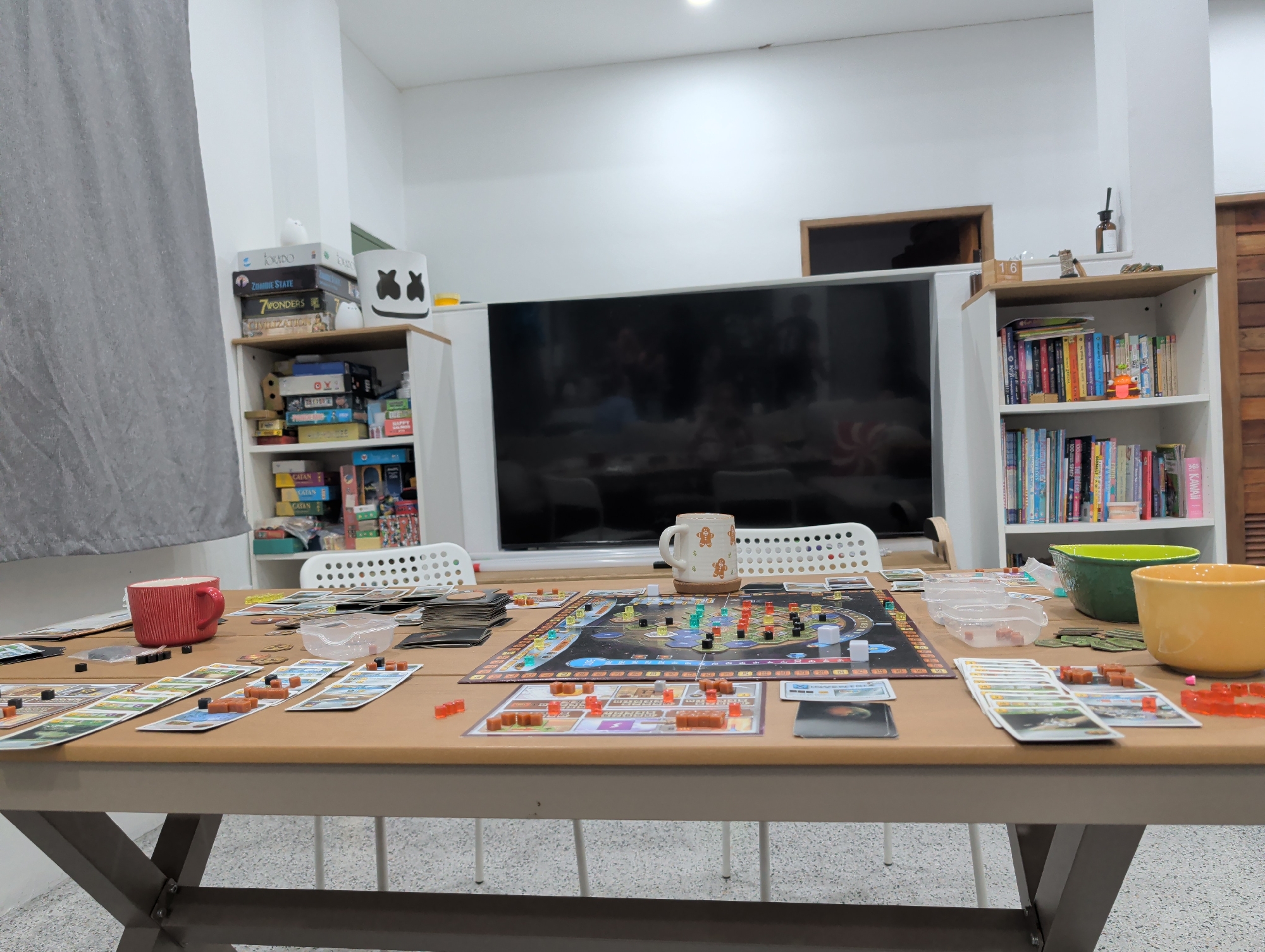 A brightly lit indoor scene shows a large table laden with the components of a board game. The game is likely "Terraforming Mars," with a central game board, player boards, cards, and various resource tokens scattered across the surface. Two mugs, one red and one decorated with gingerbread men, are also on the table. Clear plastic containers hold game components. White chairs are positioned around the table, indicating the potential presence of players. The backdrop features a modern interior with white walls, shelves stocked with books and games, and a large television. The image does not provide any specific information linking it to Thong Sala, Thailand.