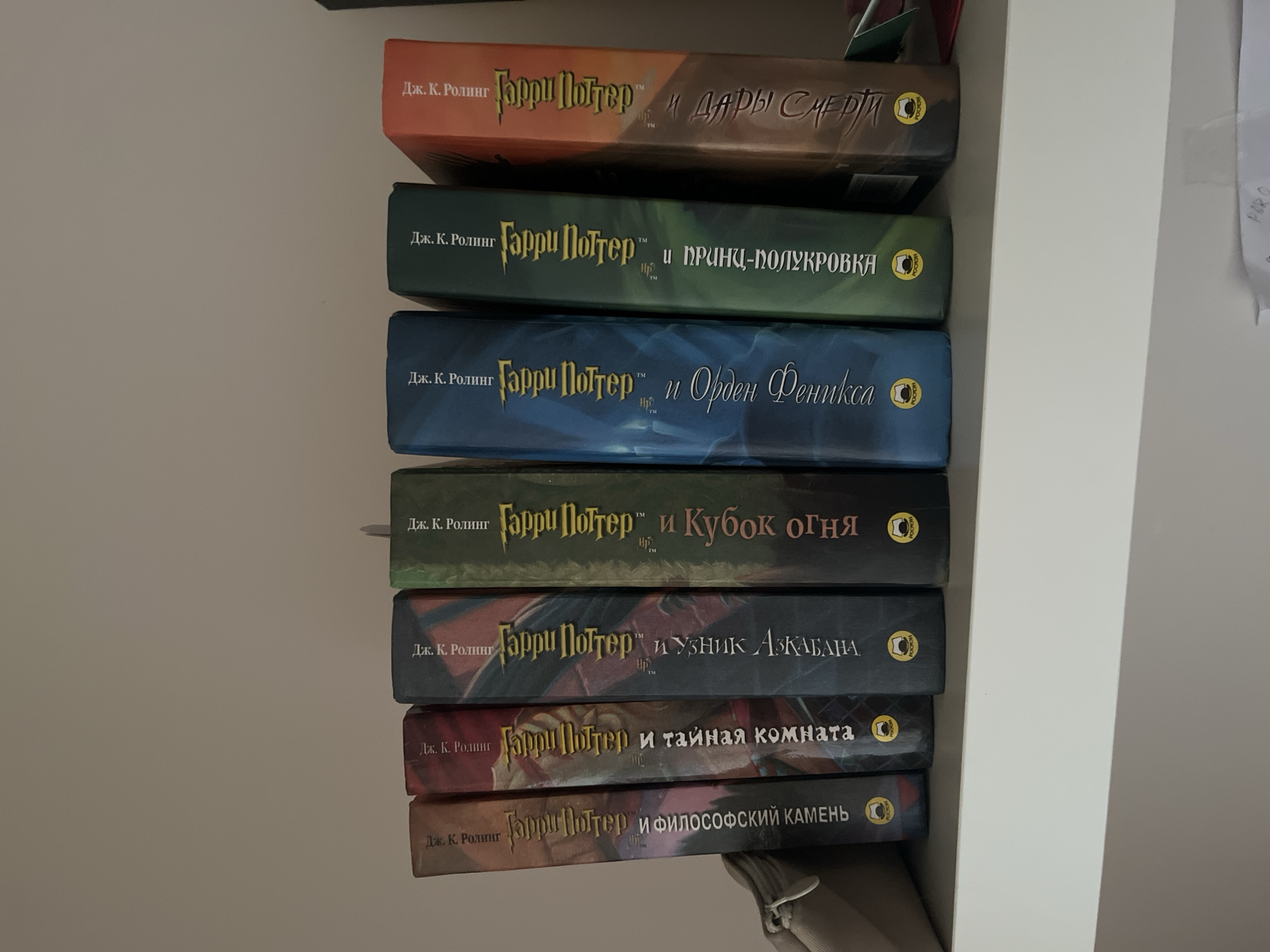 Seven Harry Potter books are displayed upright on a white shelf. The books are arranged from left to right, with their spines facing the viewer. Each book has the title "Гарри Поттер" (Harry Potter) printed vertically in yellow lettering, along with the author's name "Дж. К. Роулинг" (J.K. Rowling) above it. The books are of various colors, including brown, red, dark blue, dark green, and orange. Some of the books have additional text in Russian below the main title. At the bottom of the spine of some books, a circular logo with a bird silhouette is visible. The shelf is situated against a plain, light-colored wall. The image appears to have been taken in a residential setting. The specific location in Haabneeme alevik, Estonia, cannot be determined from the image alone.