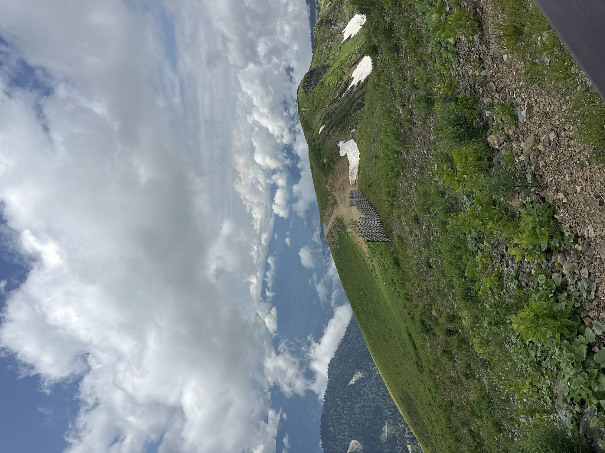 The image depicts a mountainous landscape in Estosadok, Russia, under a cloudy sky. The foreground is dominated by a rocky, sparsely vegetated slope with green plants and grass. A rustic wooden fence runs along a winding dirt path that ascends the mountain. In the mid-ground, a lush green mountainside slopes upwards, with patches of snow visible near the peak. Further in the distance, layers of mountains are shrouded in clouds, creating a sense of depth. The overall scene conveys a natural, rugged environment, likely a popular hiking or sightseeing spot.
