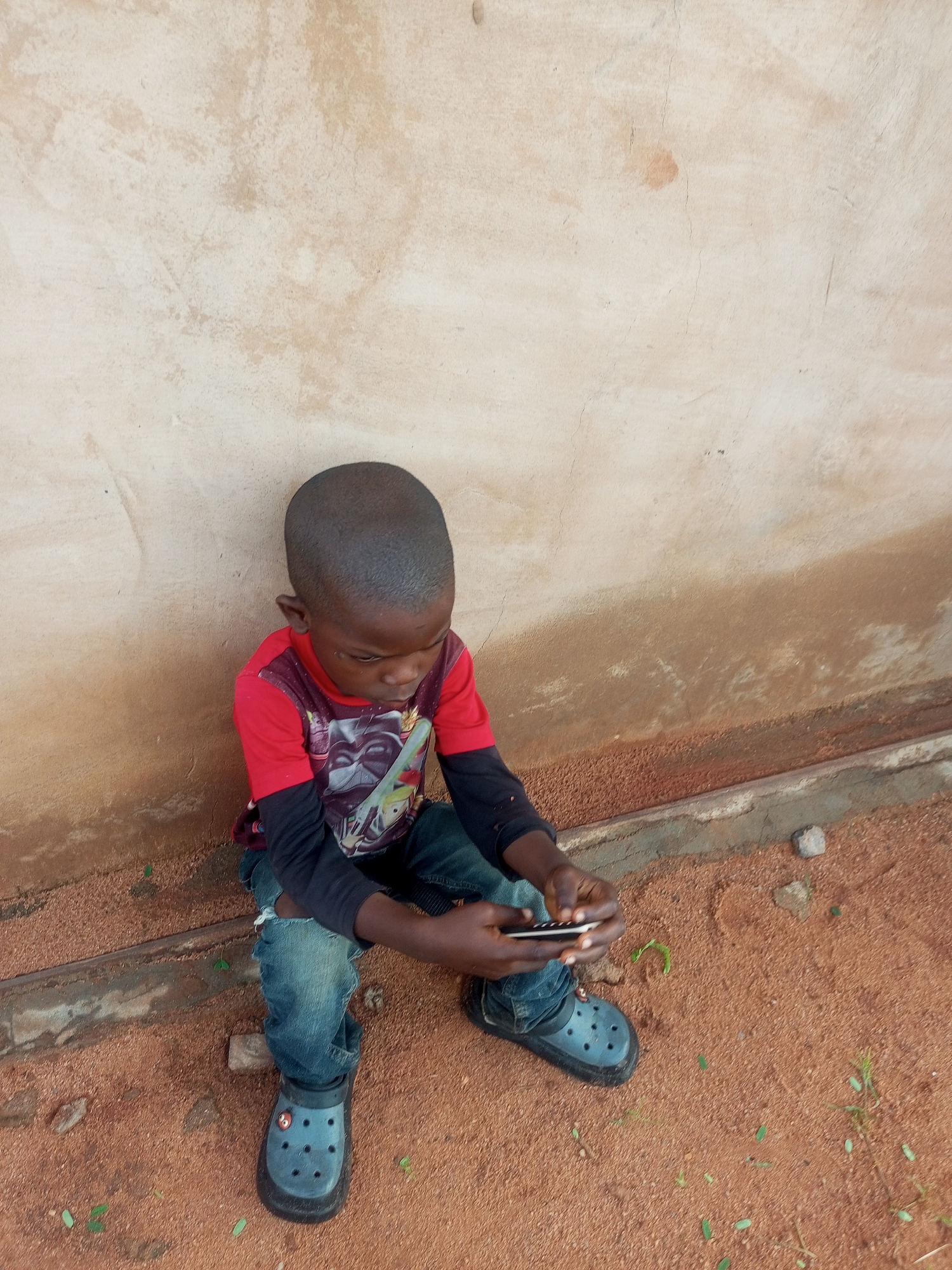 A young boy with dark skin and a shaved head sits on the ground in front of a weathered, light-colored wall. He is wearing a red t-shirt with a black long-sleeved shirt underneath, blue jeans with a rip in the left knee, and black Crocs-style shoes. The boy is looking down at a smartphone he is holding in his hands. The ground is covered in red dirt with some small stones and green sprigs of vegetation. The wall behind him has an uneven texture and color variations, with patches of brown and lighter shades. The scene appears to be outdoors, in what could be a rural or less developed urban setting. There is no indication of the specific location within Tempure, Nigeria, other than that it is a place.