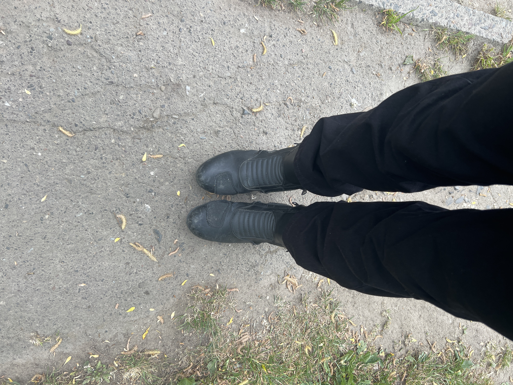 The image shows a person's legs and feet standing on a cracked concrete surface in Cherliany, Ukraine. The person is wearing black pants and black leather motorcycle boots. The concrete is light gray and has small cracks and patches of discoloration. There are scattered dead twigs and dry grass visible on the concrete and along the edges of the frame. A patch of green grass is visible along the top and right edges of the frame.
