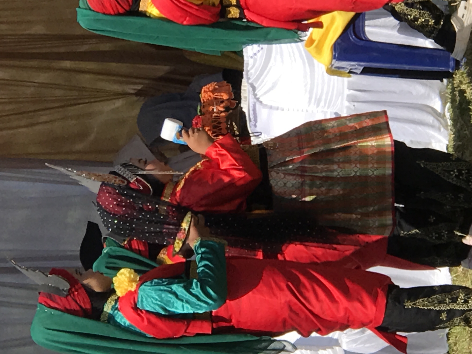 In Pantonrayeuk Satu, Indonesia, three individuals in traditional attire are positioned outdoors. The person on the left wears a red sash over a teal garment, a red headscarf, and black pants with gold embroidery. To their right, another individual is adorned in a black and red polka-dot head covering, a red top, and a skirt with woven gold and orange patterns, also with black embroidered pants. This person holds a white cylindrical object and a string of orange decorative items. The third individual on the far right is partially visible, dressed in a red top and green headscarf, with a yellow garment draped over their shoulder. They are wearing black pants with gold embroidery. Behind them, a brown curtain hangs, and a white draped table is visible, along with a blue chair. The ground is covered in grass.