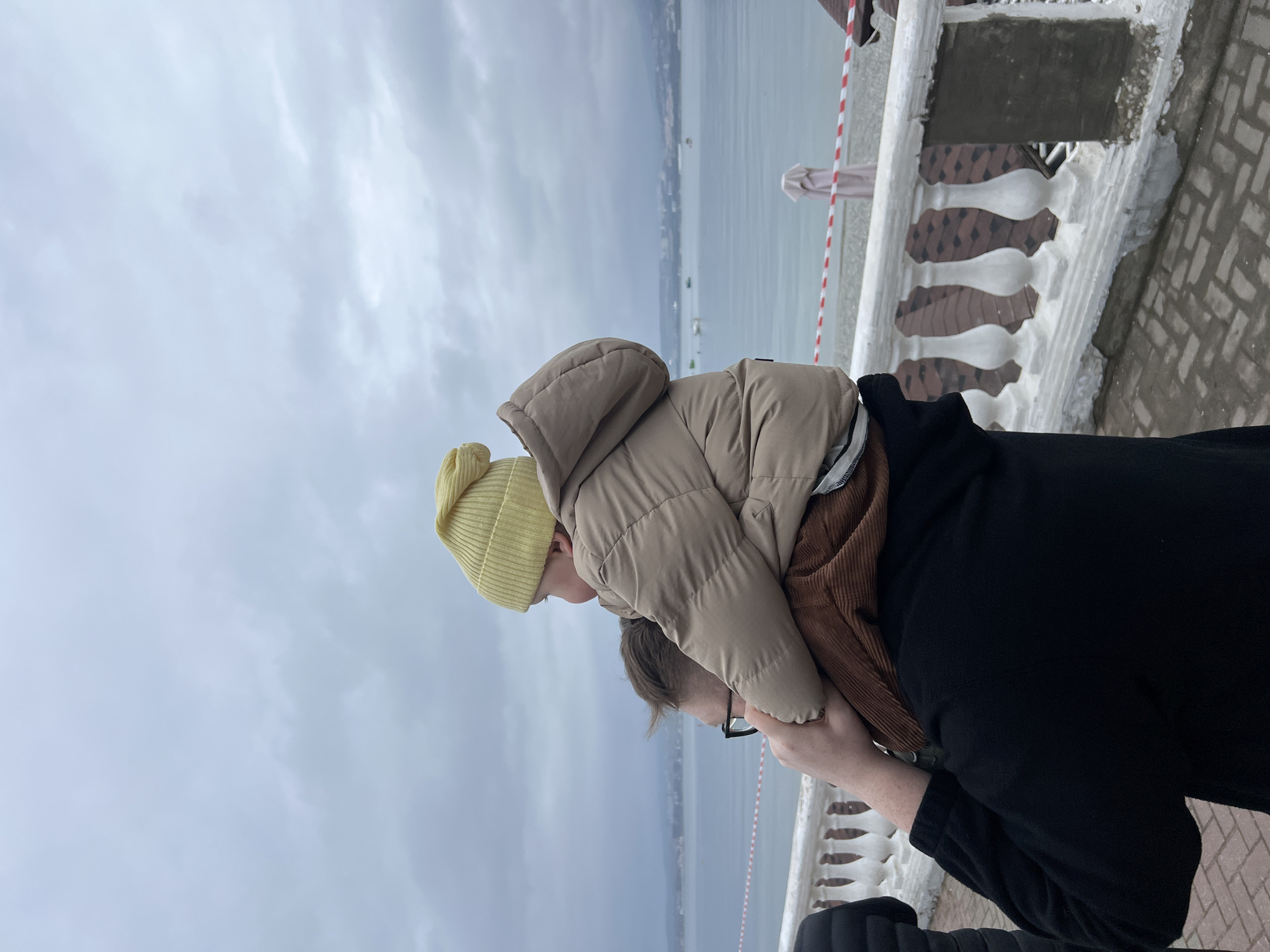 The image captures an outdoor scene in Gelendzhik, Russia, under an overcast sky. In the foreground, an adult is visible from the back and left side, wearing a black long-sleeved garment and dark-rimmed glasses. This adult is carrying a small child on their shoulders. The child, oriented away from the camera and looking towards the water, wears a yellow knitted beanie and a beige padded winter jacket. Brown corduroy fabric is visible on the child's lower body and also on the adult's shoulders, suggesting either matching attire or the child's lower garment draped over the adult. The adult's hands are visible gripping the child's legs/thighs.
The ground is paved with reddish-brown interlocking pavers. A white concrete balustrade with ornate balusters separates the paved area from a lower level. Evidence of concrete patch repair is visible on sections of the balustrade. Below the balustrade lies a light-colored pebbly beach, where a red and white striped warning tape or barrier is stretched, and a folded light-colored beach umbrella stands. Beyond the beach is the calm, gray-blue water of Gelendzhik Bay. In the distance across the bay, a hilly or mountainous shoreline is visible, dotted with scattered buildings and several small boats on the water. The sky is uniformly gray with cloud cover.