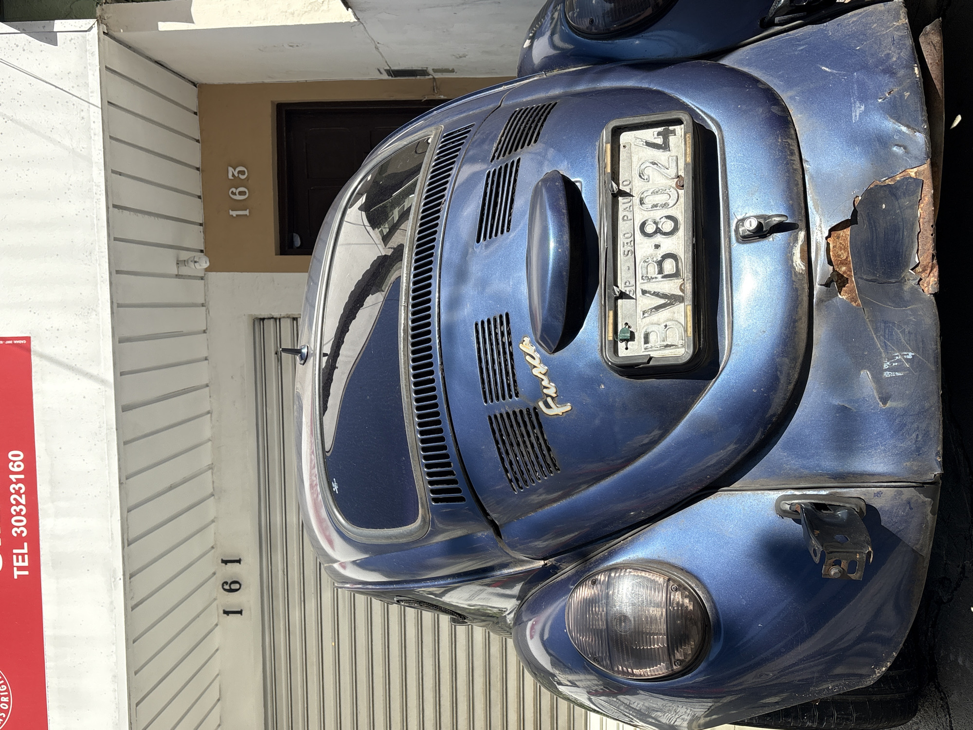 The image shows the rear of a blue Volkswagen Beetle parked in front of a building in São Paulo, Brazil. The car has a license plate that reads "BVB-8024" and "SP - SAO PAULO". The building has numbers "161" and "163" on its facade. The car appears to be damaged on the lower rear bumper. The background shows a roll-up garage door and a dark-colored door. The scene is illuminated by sunlight, casting shadows.