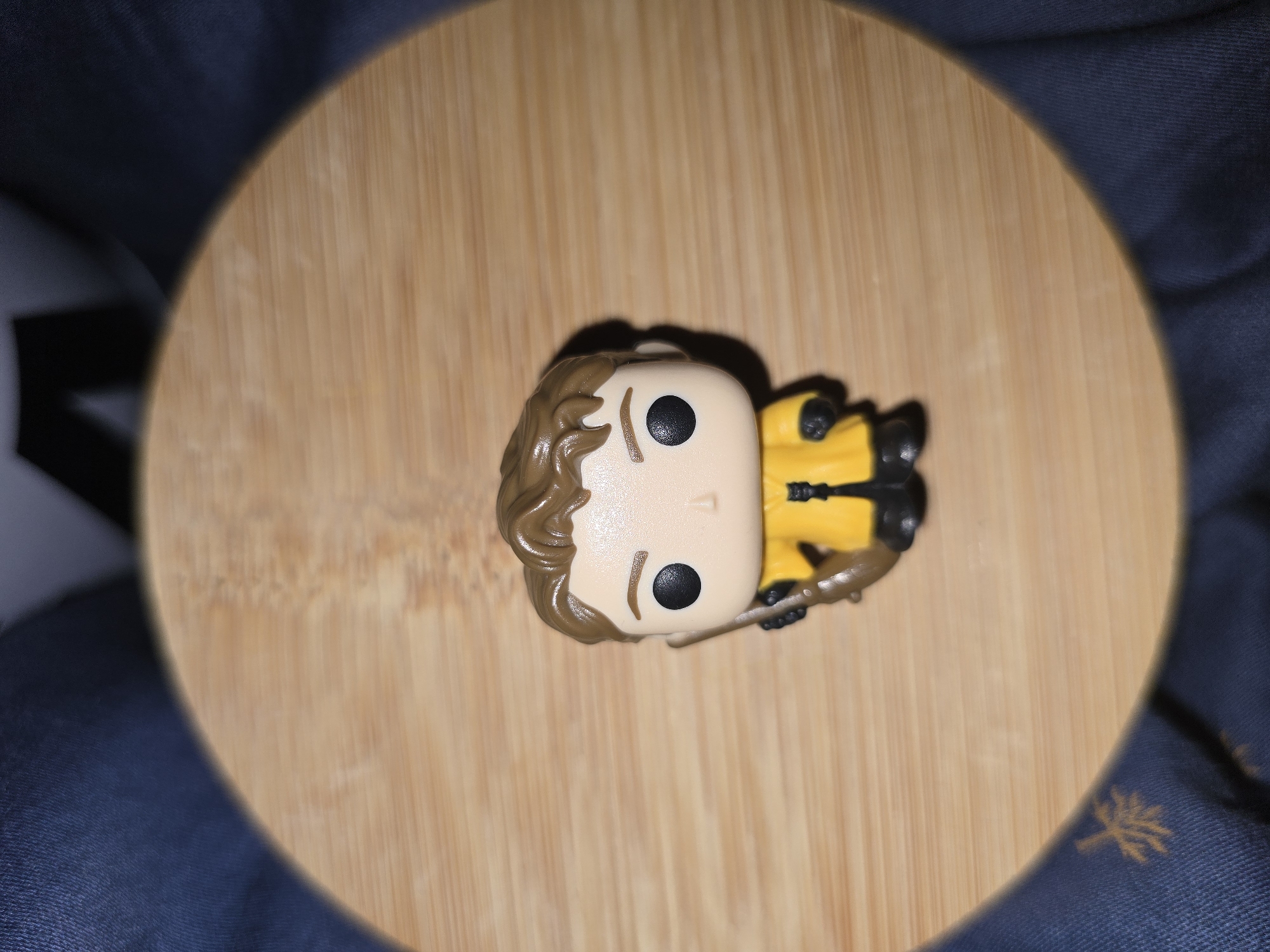 The image shows a Funko Pop figurine of Harry Potter in his Quidditch uniform, lying on its back on a wooden surface. The figurine is primarily yellow and black, with brown hair and black eyes. It appears to be holding a broomstick. The background is a dark blue, textured fabric, possibly a blanket or clothing, with a subtle tree-like pattern visible on the right side of the image. The scene is lit by a light source that casts a shadow from the figurine, creating a vignette effect with a bright, circular area focused on the figurine, fading to darkness at the edges. There is no visible text. The overall impression is of a close-up shot of a collectible toy. The location is Petropavlivska Borshchahivka, Ukraine.