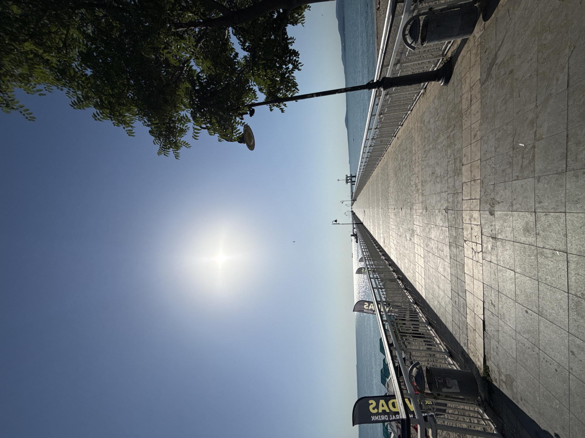 The image shows a perspective view down a long, paved pier extending over the sea. The pier is lined with a metal railing and several lampposts. On the left side of the pier, there are flags with black and yellow designs, beach chairs, and a trash bin. The sea is visible to the left of the pier, with distant hills on the horizon. The sun is bright in the clear blue sky, casting long shadows on the pier. The scene appears to be set in Burgas, Bulgaria, likely on the Burgas Pier, a popular promenade. There are no people visible in the immediate foreground, though a few figures can be seen further down the pier and on the beach to the right.