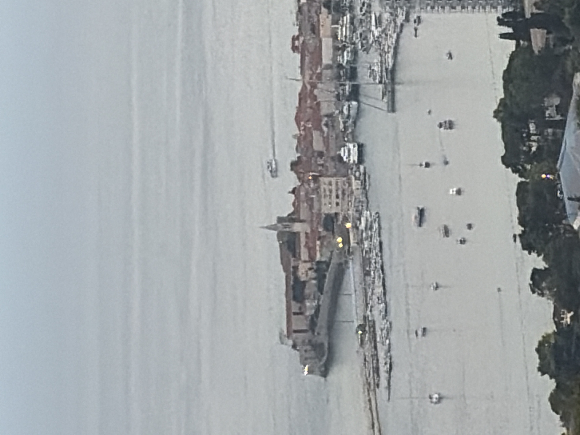 This aerial image depicts a coastal city with a fortified old town situated on a peninsula. The old town, characterized by its stone walls and red-tiled roofs, features a prominent church spire. A marina, filled with numerous boats of various sizes, is located adjacent to the old town. Several vessels are also visible in the open water, with one ferry-like boat in the distance. The overall scene is one of a peaceful maritime city, possibly during daylight or twilight hours, given the muted lighting. The location is identified as Budva, Montenegro.