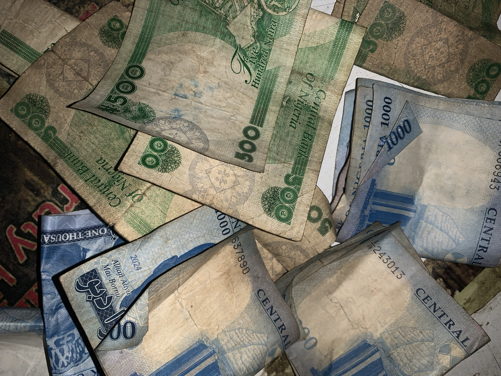 The image displays a disordered pile of Nigerian Naira currency notes. The denominations visible include 500 Naira (green notes) and 1000 Naira (blue notes). The notes appear used and somewhat soiled. Some notes are partially visible, with serial numbers and inscriptions of "Central Bank of Nigeria" and "One Thousand" or "Five Hundred Naira" discernible. There is no indication of the location of the scene in New Stadium, Nigeria or the presence of people.