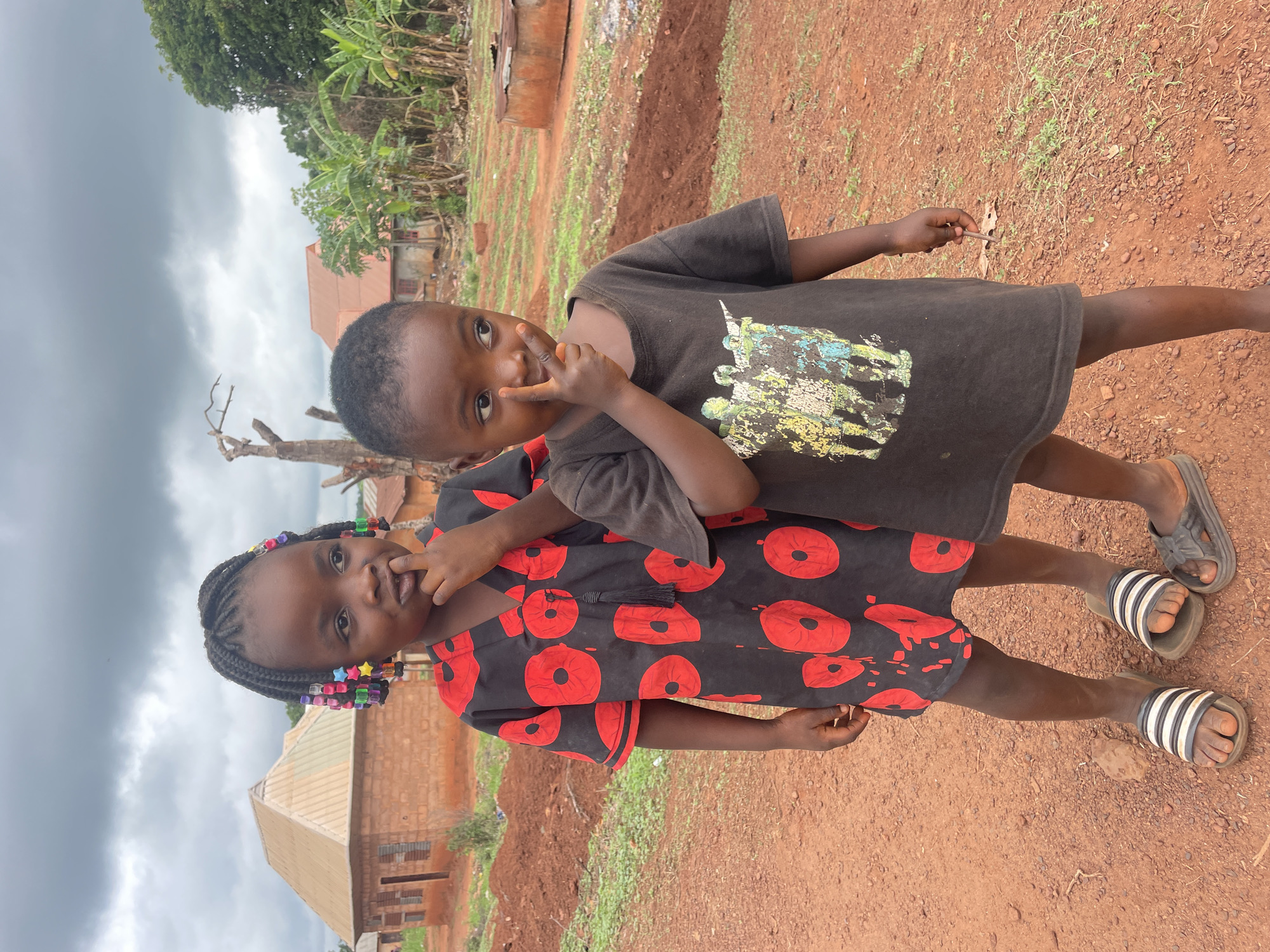 Two children stand outside in Ogbe, Nigeria. One child, a girl, wears a black dress with red circles on it and has colorful beads in her braided hair. She wears striped sandals. The other child, a boy, wears a black shirt with a graphic print. He wears dark sandals. Both children are touching their faces with their hands. The ground is red clay. In the background are houses and trees. The sky is overcast.