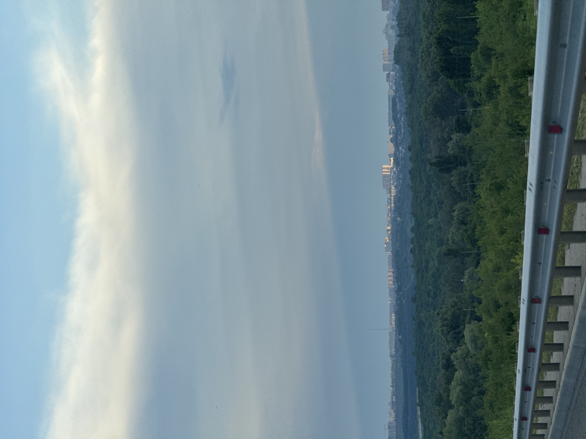 The image depicts a panoramic view of a city skyline in Novomar'evskaya, Russia, seen from a slightly elevated vantage point. In the foreground, a silver metal guardrail with red reflectors runs horizontally across the lower portion of the frame, suggesting the perspective is from a road or elevated pathway. Beyond the guardrail, a dense expanse of lush green trees covers the mid-ground, forming a natural buffer between the viewer and the urban landscape.
The city skyline, located in the distance, is characterized by a collection of multi-story buildings, including apartment blocks and other urban structures. The buildings are illuminated by soft, warm light, indicating either dawn or dusk. A tall, slender structure, possibly a radio tower or a communication mast, stands prominently amongst the cityscape.
The sky above is a mix of pale blue and diffused white clouds. A large, bright white cloud formation stretches across the upper portion of the image, with lighter wisps and streaks of white scattering across the pale blue expanse. A faint, horizontal band of shadow or darker sky is visible in the mid-sky, possibly cast by a cloud formation not entirely within the frame. The overall atmosphere of the image is serene and suggests a peaceful, early morning or late evening observation of the city. There are no people or visible animals in the frame.