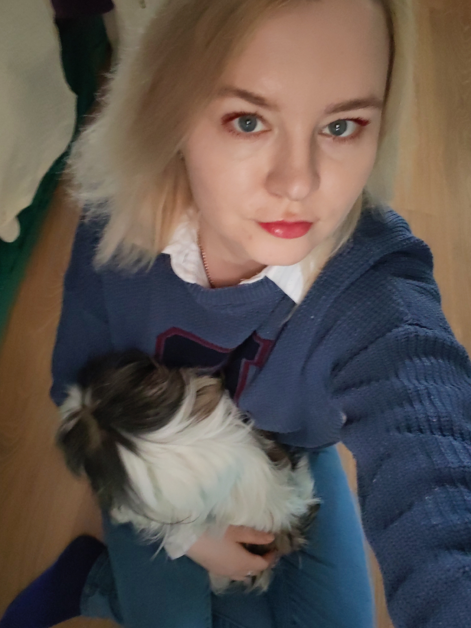 A blonde woman with fair skin is holding a small dog in her lap. The woman is wearing a blue sweater over a white collared shirt and blue jeans. She has red eyeliner and lipstick. The dog is black and white, and it is looking towards the left. The background is a wooden floor. The location cannot be determined to be Izhevsk, Russia, based on the provided image.