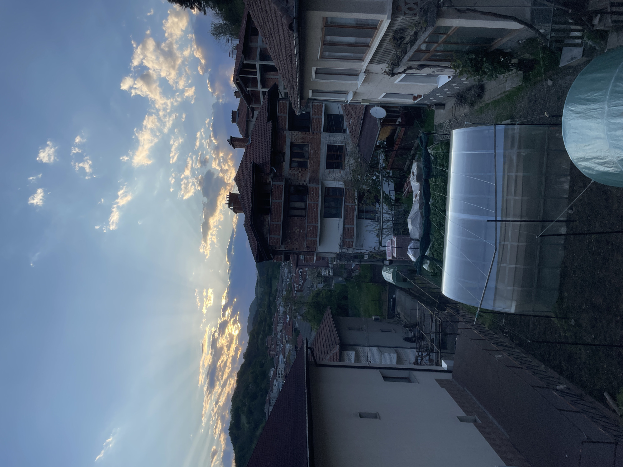 The image shows a view from an elevated position in Zlatograd, Bulgaria, at what appears to be dusk or dawn. The sky is visible, with scattered clouds illuminated by a low sun or setting sun, creating a play of light and shadows. The sun's rays are piercing through the clouds, adding a dramatic effect.
In the foreground, there are residential buildings of varying designs and construction stages. Some buildings are brick, while others have finished facades. There are tiled roofs and windows visible on the structures.
A greenhouse-like structure is visible on the right side of the frame, with a translucent covering, suggesting its use for growing plants. Beyond the buildings, the landscape descends, revealing a town nestled among hills or mountains, giving the impression of a valley setting. The overall environment appears to be a residential area on the outskirts of a town, with a mix of construction, dwellings, and nature. No people are visible in the image.