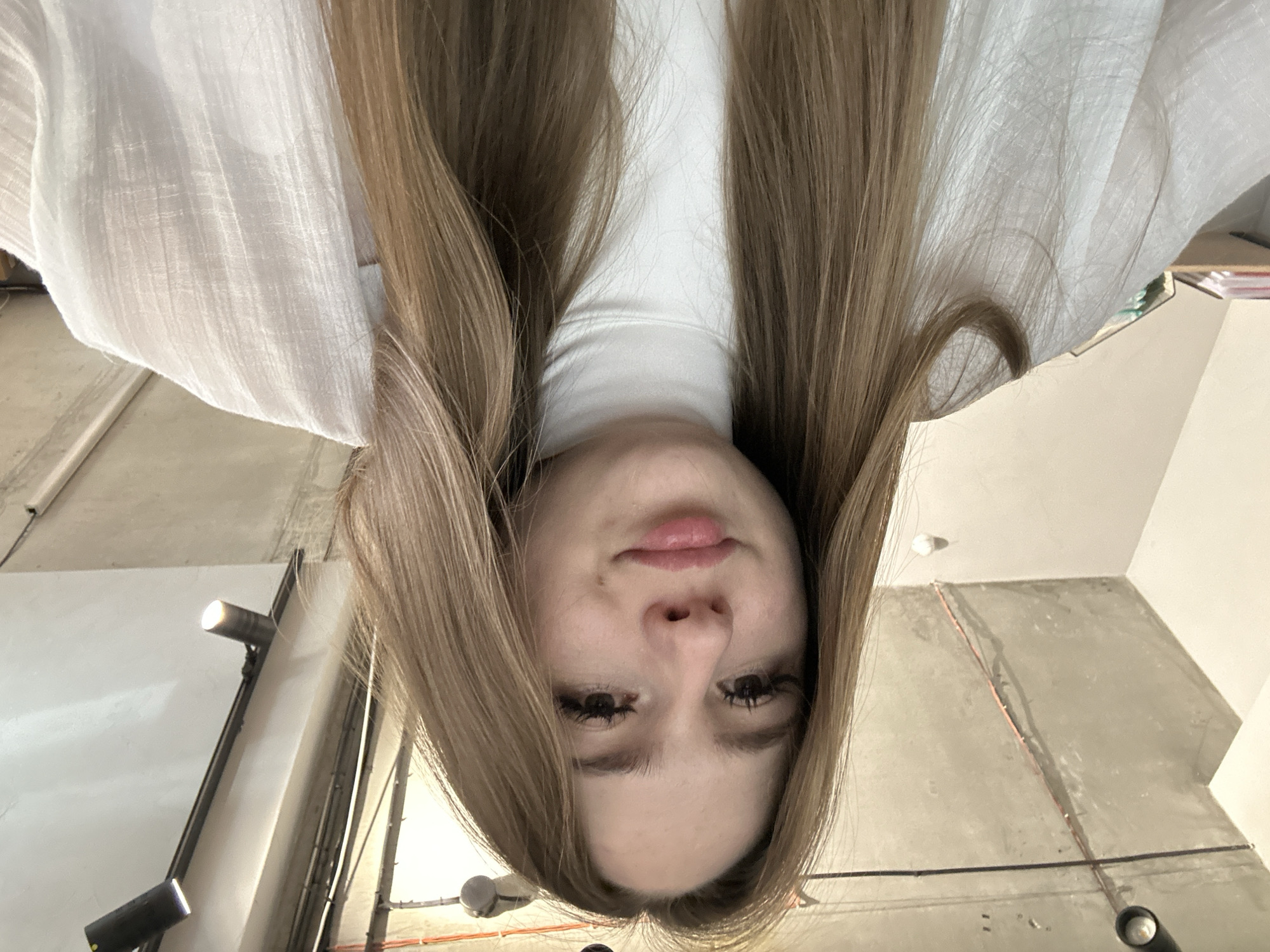 A young woman with long, straight blonde hair and dark brown eyes looks directly at the camera, her mouth in a slight pout. She is wearing a white turtleneck sweater and an unbuttoned white shirt over it. The background shows a concrete ceiling with exposed pipes and track lighting, suggesting an urban, industrial-style interior, possibly a cafe or co-working space. The lighting is bright, casting soft shadows.