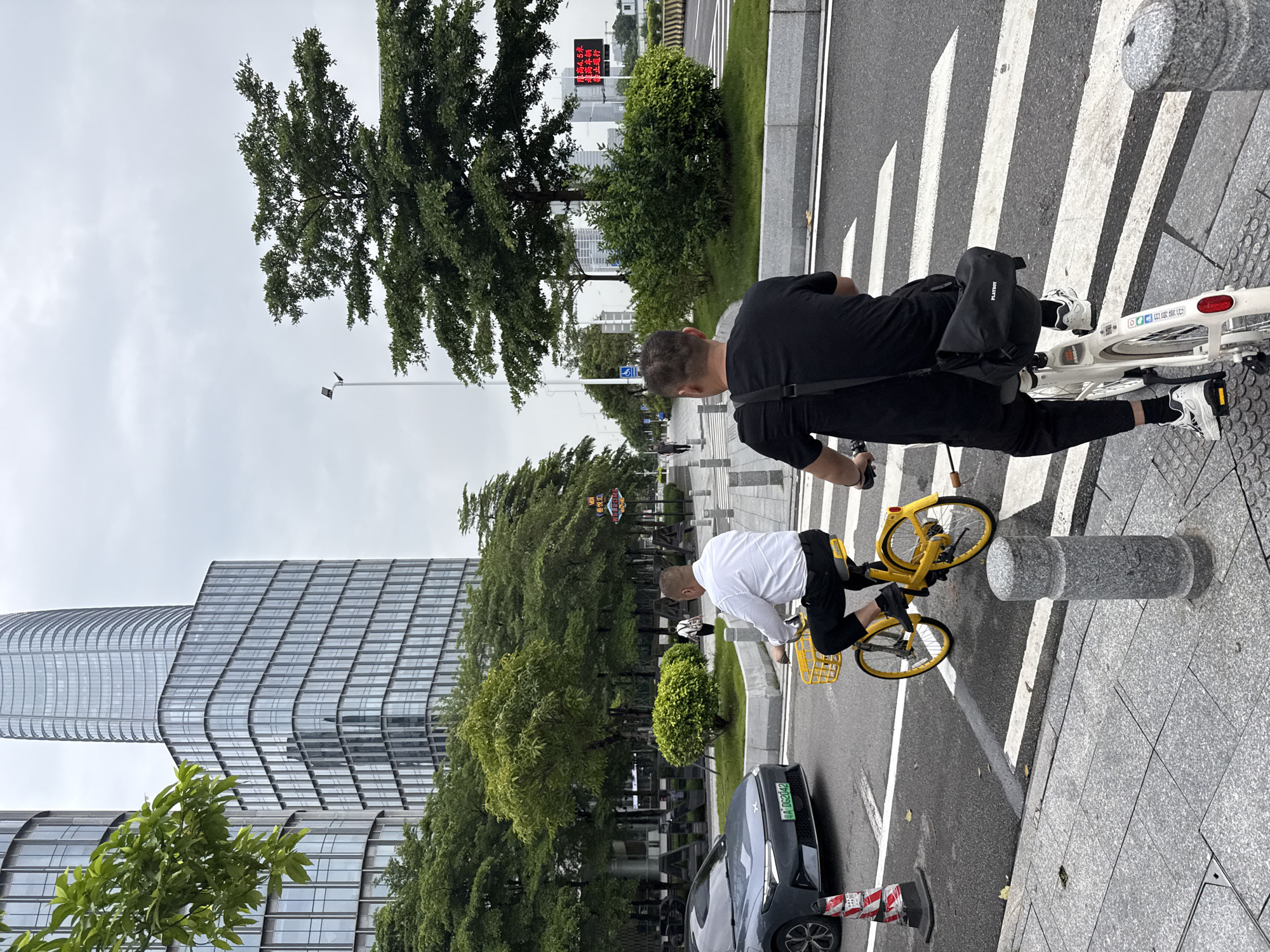 Two men are riding bicycles on a street in Haizhu District, China. The man in the foreground is riding a white bicycle and is wearing a black t-shirt, black pants, and white sneakers. He has a black bag slung over his shoulder. The man in the background is riding a yellow bicycle with a yellow basket and is wearing a white shirt and black pants. They are both on a street with a crosswalk. In the background, there are modern glass buildings and trees. The sky is overcast. A gray car is stopped at the crosswalk.