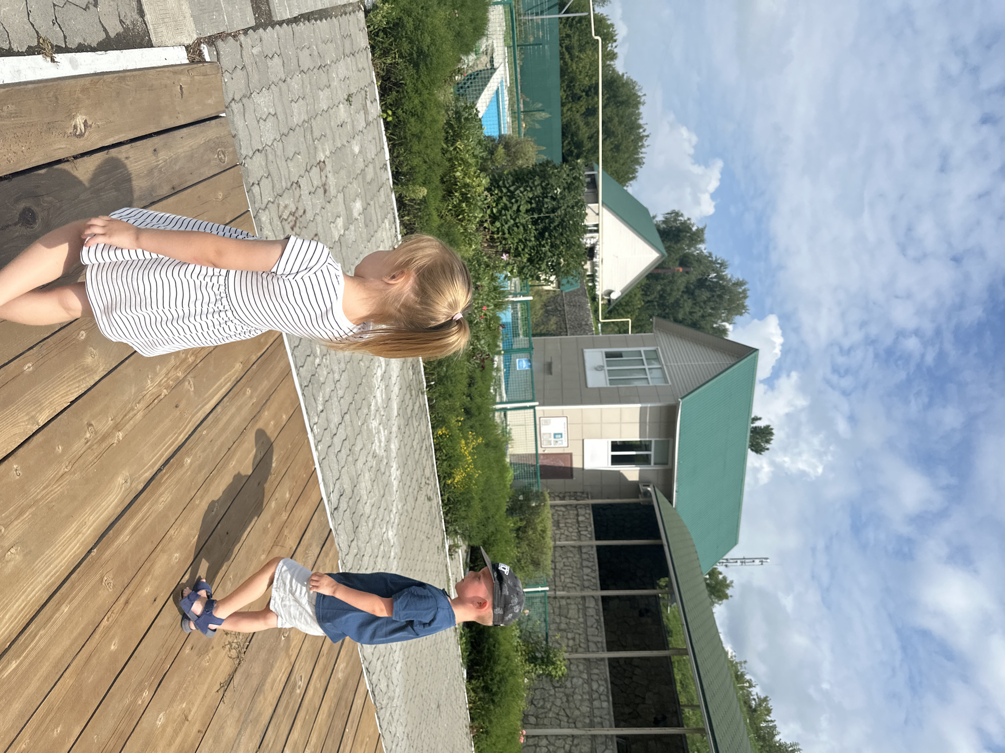 The image depicts an outdoor scene with two children, a girl and a boy, in the foreground. The girl, facing away from the camera and towards the left, has blonde hair in a ponytail and is wearing a black and white striped dress. The boy, to the right and also facing away, is wearing a blue t-shirt, white shorts, a grey baseball cap, and blue sandals. They are standing on a wooden platform.
In the background, there are several buildings with green roofs, surrounded by lush greenery and trees. A swimming pool with bright blue water is visible in the middle ground, enclosed by a fence. The ground is paved with gray interlocking stones. The sky is partly cloudy with a mix of blue sky and white clouds. The overall setting appears to be a recreational area or a resort, possibly in or near the city of Danilovka (Altai), Russia, given the context. The lighting suggests it is a sunny day.