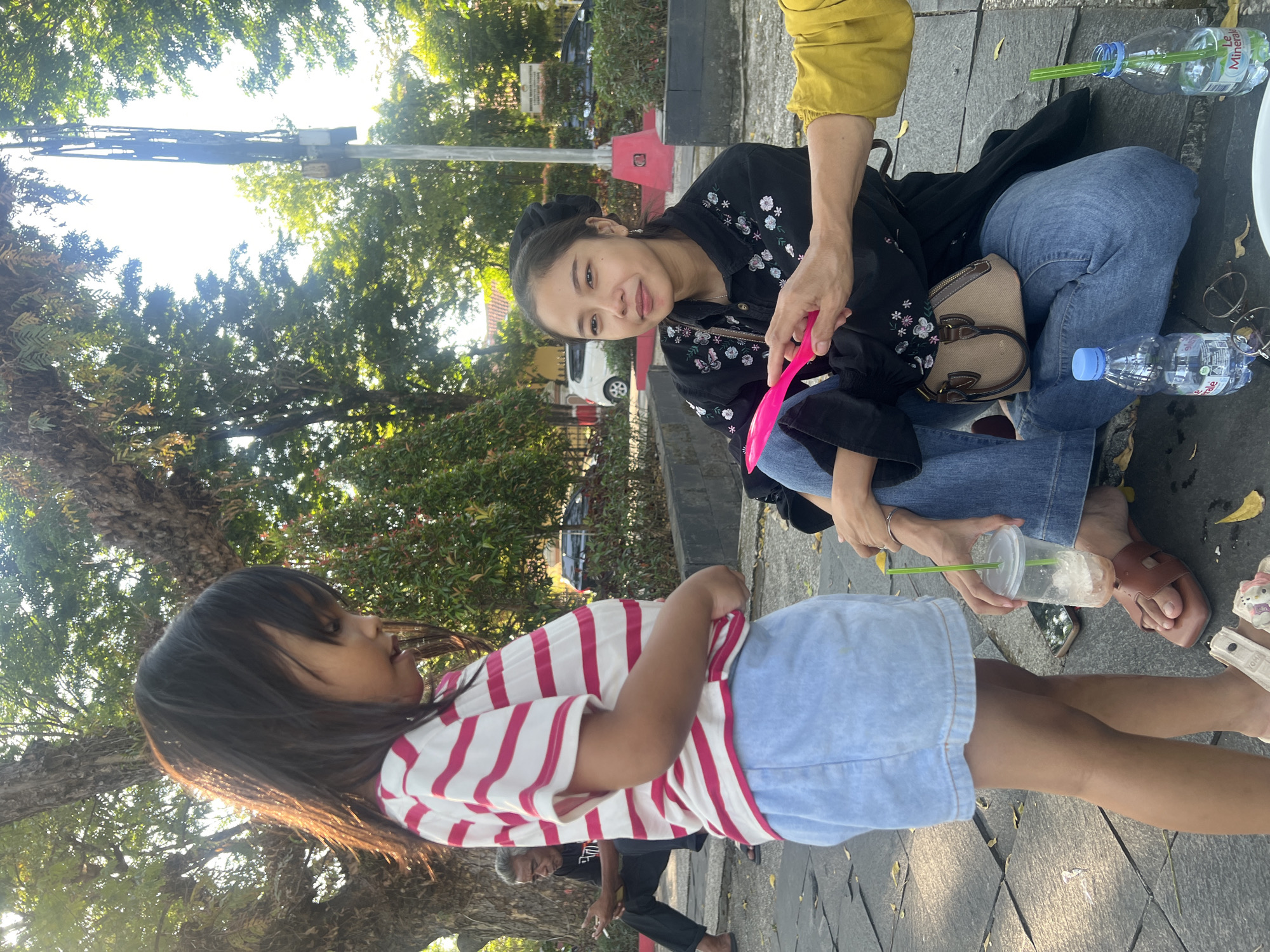 In Purwodadi, Indonesia, a young girl in a red and white striped shirt and denim shorts stands outdoors, facing a woman who is sitting cross-legged on the ground. The woman, wearing a black shirt with floral embroidery and blue jeans, is holding a pink plastic utensil and looking at the girl. A third person, wearing a yellow top, is partially visible to the right, also interacting with the woman. The ground is paved with stone slabs, and fallen leaves are scattered around. In the background, trees with dense foliage create a shaded environment, and a tall metal tower can be seen in the distance. Two plastic water bottles, one unopened and one with a green straw, are placed on the ground near the sitting individuals. An elderly man is visible in the far background, seated on a ledge and looking at his phone. The scene appears to be a public outdoor area, possibly a park or plaza.
