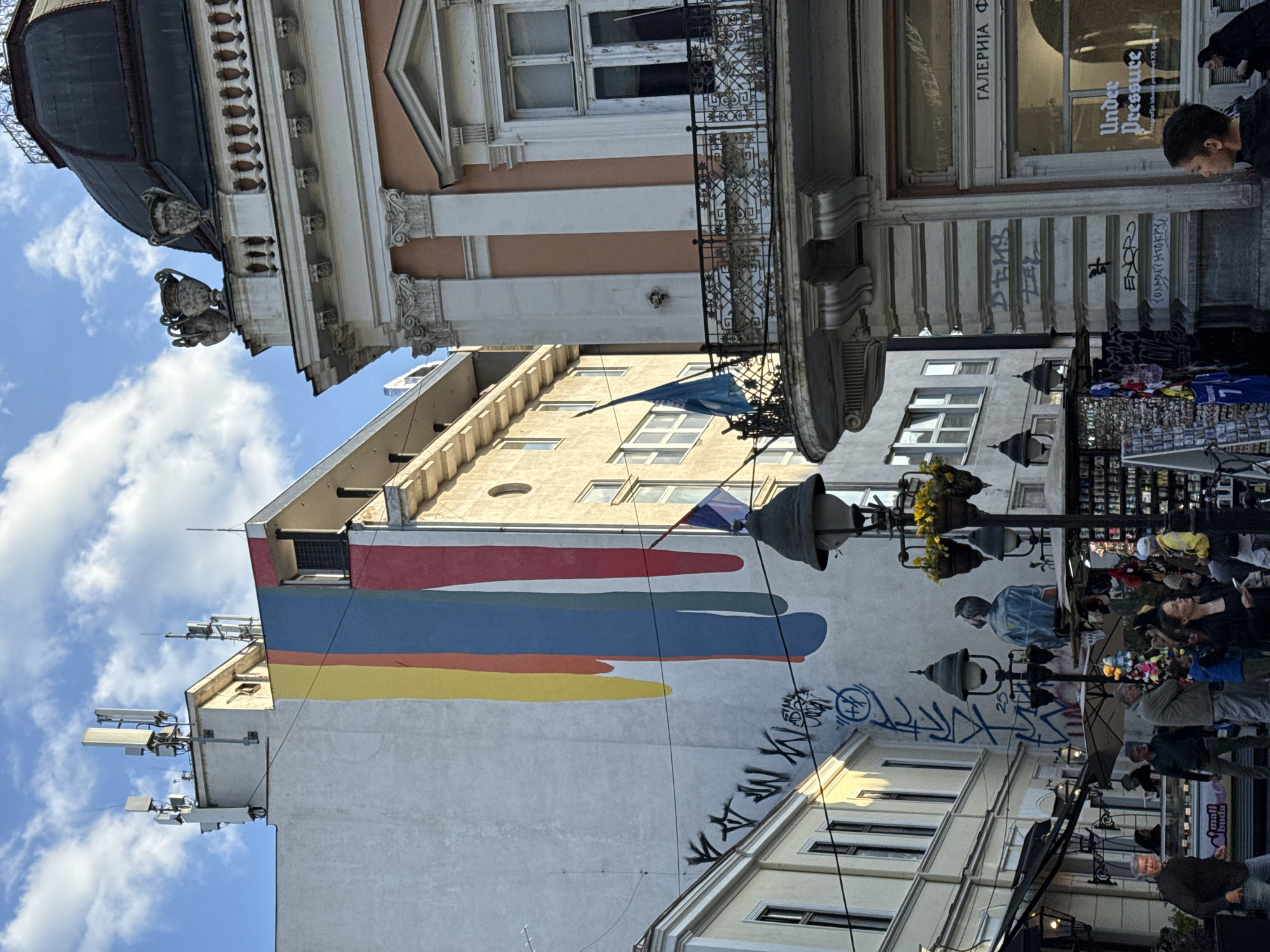 This media file captures a vibrant street scene in Belgrade, Serbia, on a bright, cloudy day. The image, oriented correctly, shows a narrow urban thoroughfare flanked by buildings of varying architectural styles.
To the left, a grand, ornate building features a dark, possibly metal, dome-like structure and decorative urns, cornices, and columns along its light-colored facade. In the center, a taller, plainer building stands out with a large mural of bold red, blue, and yellow horizontal brushstrokes. Below this, a stylized painting of a man is visible. The rooftop of this building is equipped with multiple cell phone antennas. Further down the street on the right, another building shows balconies and windows.
The street level is bustling with activity. Numerous people are seen walking, standing, and browsing merchandise at what appear to be street vendors or market stalls displaying items like clothing and small colorful toys. Several ornate street lights, some adorned with hanging flower baskets, illuminate the scene. Graffiti and tags are prevalent on the lower sections of the buildings.
Visible text includes: "ГАЛЕРИЈА Ф" (GALERIJA F - Gallery F), "Under Pressure", "grafija izbor", "buda", "EXOPO", and various graffiti tags such as "DEMS", "ZEL", and "OYOVL YAMARRY". A small tri-color flag (possibly Serbian) hangs from one building, and a larger blue flag from another. The overall impression is that of a lively commercial or pedestrian zone in a historic European city.