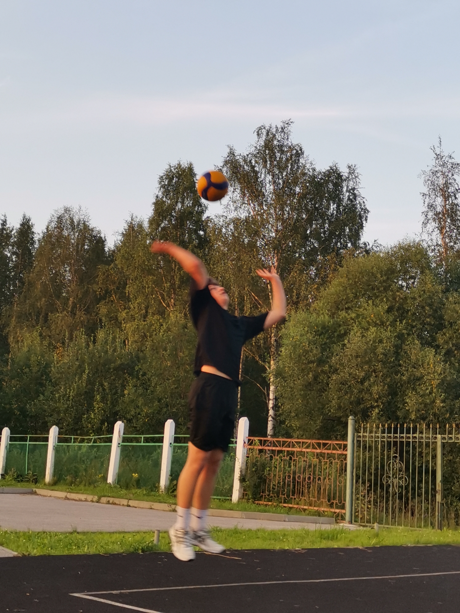 A young male is playing volleyball in an outdoor setting, captured mid-jump as he prepares to hit the ball. He is wearing a black t-shirt and black shorts, with white socks and white athletic shoes. The volleyball, orange and blue, is suspended in the air above him.
The background consists of a dense line of green trees under a pale sky. The setting appears to be a sports ground or park, with a black track in the foreground. Behind the player, there's a white fence with vertical posts and a green mesh covering, separating the track from a grassy area. Further back, a decorative metal fence, partially rusted, runs horizontally, suggesting a boundary to a property or perhaps an older section of the park. The lighting indicates it is late afternoon or early evening, with the sun casting warm light and long shadows, typical of a clear, pleasant day. The overall atmosphere is one of active recreation and enjoyment of the outdoors. No visible text is present in the image.