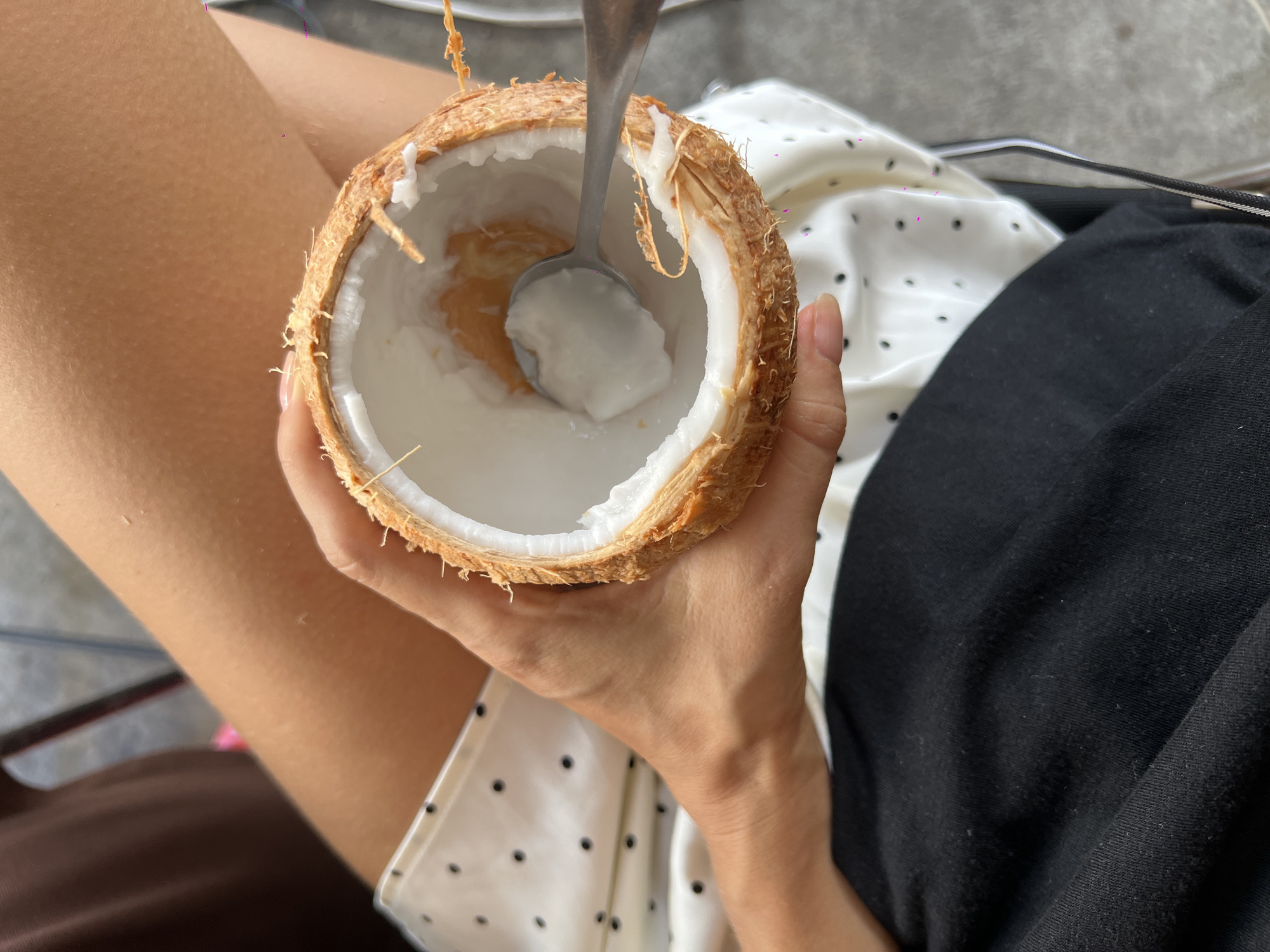 A person's hands are holding a halved coconut with a spoon inside, scraping the white flesh. The person is wearing a white dress with black polka dots and a black garment underneath. The scene appears to be outdoors, possibly in a city like Nha Trang Ward, Vietnam, indicated by the textured grey surface in the background and the presence of metal chair legs. The lighting suggests it is daytime.