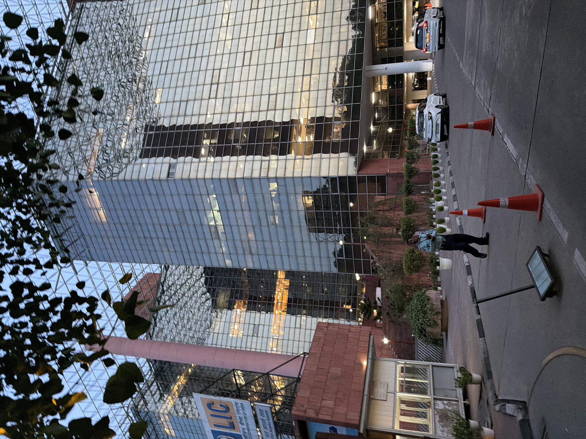 This media file depicts an exterior view of a modern glass-facade building, likely an office building, in New Delhi, India. The building's reflective surface mirrors the sky and surrounding elements, creating a dynamic visual effect. In the foreground, foliage from a tree partially obscures the view of the building.
On the right side of the image, a paved area with traffic cones suggests a road or parking lot. Two white cars are parked, and a security guard is visible standing near them, dressed in a light blue shirt and dark pants. The time of day appears to be dusk or early evening, as evidenced by the muted lighting and the sky's color. The overall atmosphere is calm and everyday, with no specific event being depicted.
Notable details include the architectural style of the building with its extensive glass panels and the presence of traffic management cones, indicating a controlled environment. The ground features a distinct black and white striped border.
Visible text includes "LIC" and some Hindi text on a banner, likely indicating an office or service associated with the Life Insurance Corporation of India. The image is captured from a slightly elevated, horizontal perspective.