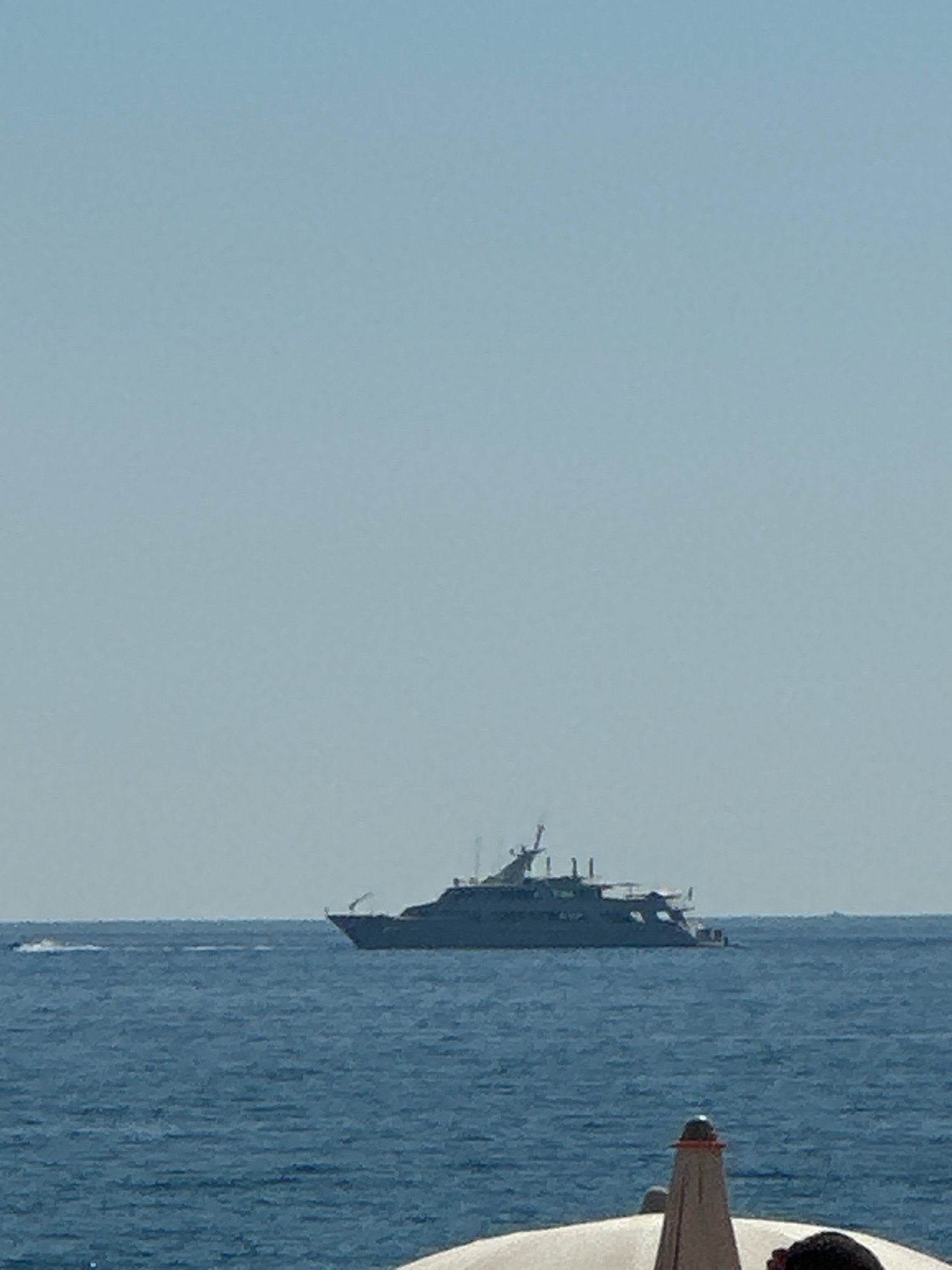 A large motor yacht is visible on the sea, under a clear blue sky. The yacht has multiple decks and appears to be at anchor or moving slowly. A smaller vessel, possibly a jet ski, is creating a wake in the distance to the left of the yacht. The foreground of the image shows a portion of a white structure, possibly a boat or a pier, with a small beacon-like object on top. The water is calm and blue. The scene suggests a leisurely day at sea.