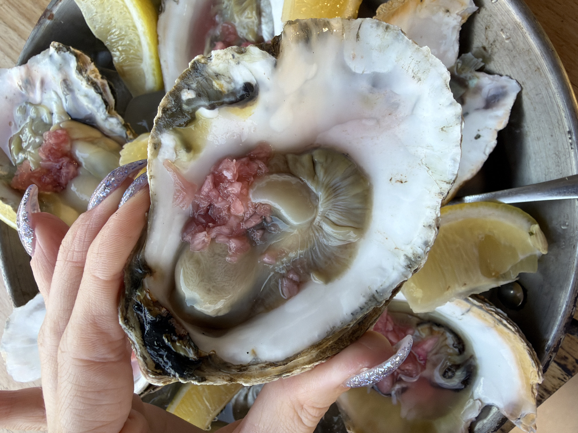 In this close-up shot, a person's hand, adorned with sparkly silver nails, holds a fresh oyster, showcasing its succulent interior. The oyster, nestled in its shell, is presented with lemon wedges and appears to be part of a larger platter of seafood. The lighting suggests a casual dining setting, possibly indoors or in a shaded outdoor area. The focus is on the texture and appearance of the oyster, hinting at a culinary experience. There are no visible text elements or explicit cues about the time of day or weather. The scene is centered around the enjoyment of fresh oysters. The background shows other oysters and lemon slices in a metal bowl, indicating a meal being served.