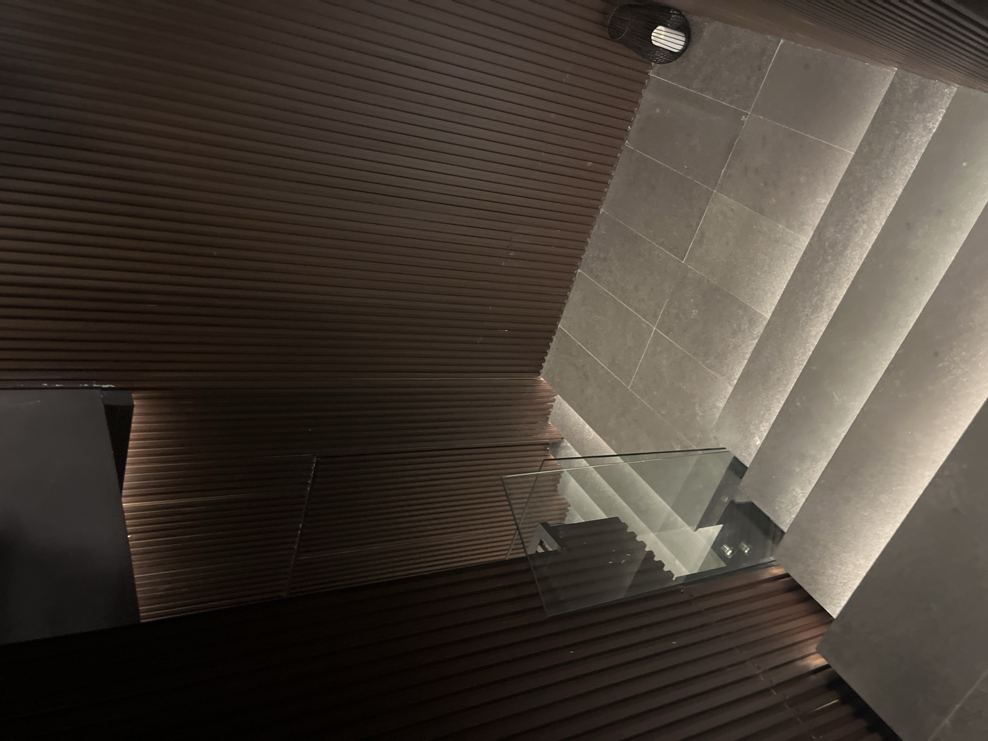 The image shows a top-down view of a modern interior featuring a staircase. The staircase is constructed with gray stone steps and a glass railing. The steps are illuminated, creating a bright contrast against the darker surroundings. The floor adjacent to the staircase is covered with brown, horizontally-lined wooden panels. The lower left of the frame reveals a dark, possibly black, rectangular surface. The upper left area shows more of the brown wooden paneling, meeting at a corner with the gray stone tiling of the staircase. A black circular object, possibly a vent or speaker, is located in the upper center of the image, where the wooden paneling and stone tiling meet. The specific location is unknown, but it's in Sofia, Bulgaria.