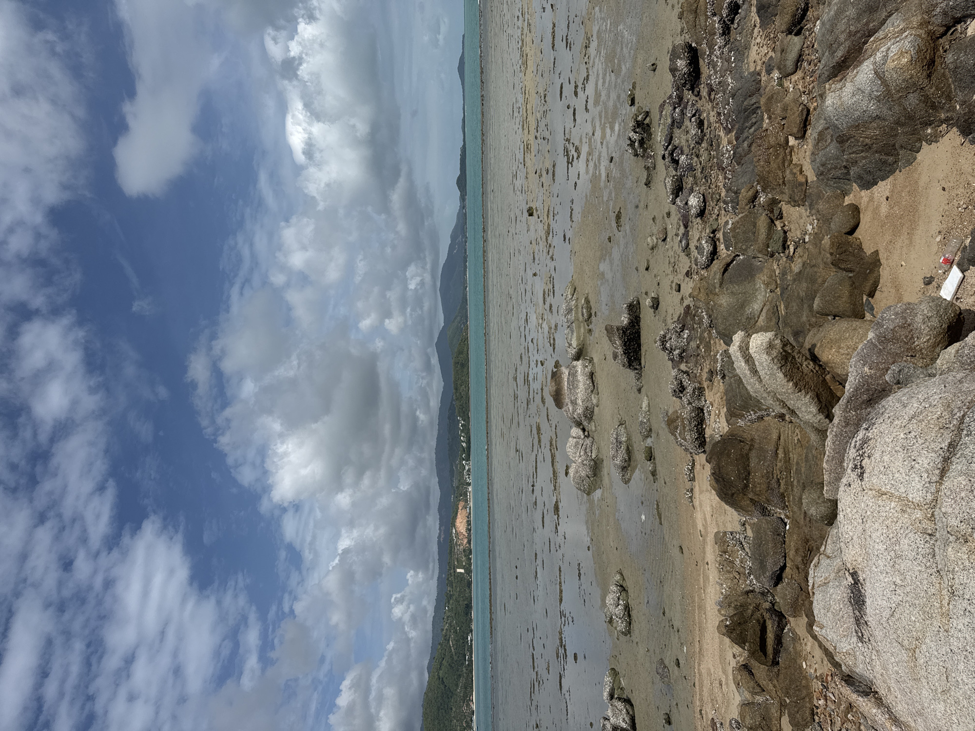 This is a vertical, medium shot of a rocky beach with a shallow sea and a distant treeline under a cloudy sky. In the foreground, large, grey and brown rocks are scattered across the sand, some of which appear to be partially submerged in shallow water. A white and red plastic bottle is visible amongst the rocks in the bottom right corner of the frame. The middle ground is a wide expanse of sand and shallow water, with smaller rocks and seaweed visible. In the background, a blue sea stretches to the horizon, meeting a distant, green treeline that extends across the width of the frame. Above the treeline, buildings and infrastructure are visible on the hillsides. The sky is a mix of blue and grey clouds, with sunlight breaking through in places. The overall impression is one of a natural, somewhat untamed coastal environment.