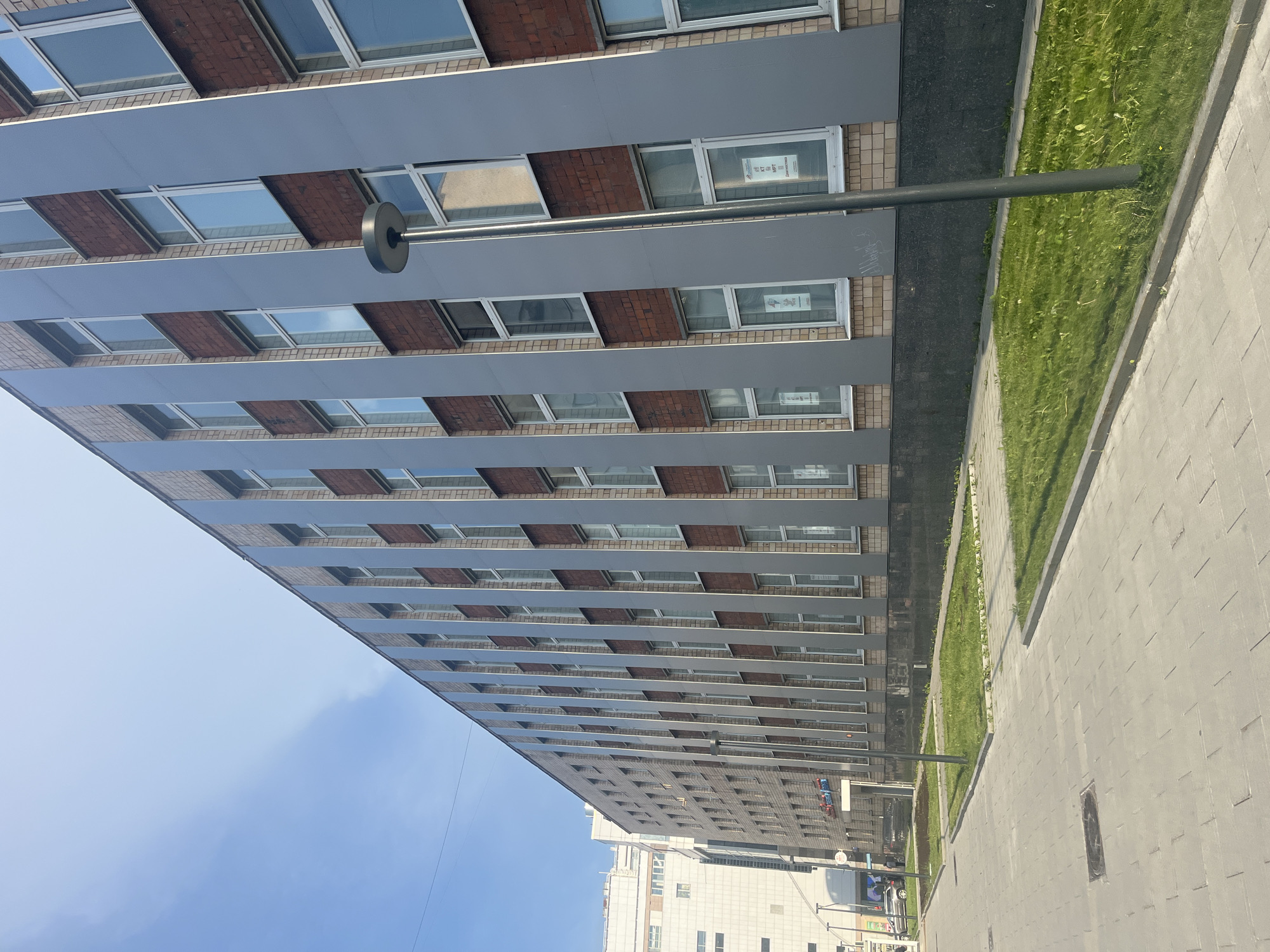 The image shows a modern office building in Moscow, Russia, on a sunny day. The building features a facade of grey metal panels interspersed with red brick accents and numerous windows. A paved walkway with grass areas runs alongside the building. A tall, slender street lamp stands in front of the building. In the background, another taller building is visible, suggesting an urban environment. The sky is clear and blue. No people are clearly visible in the immediate foreground or interacting with the building or its surroundings. The scene appears to be a typical urban streetscape.