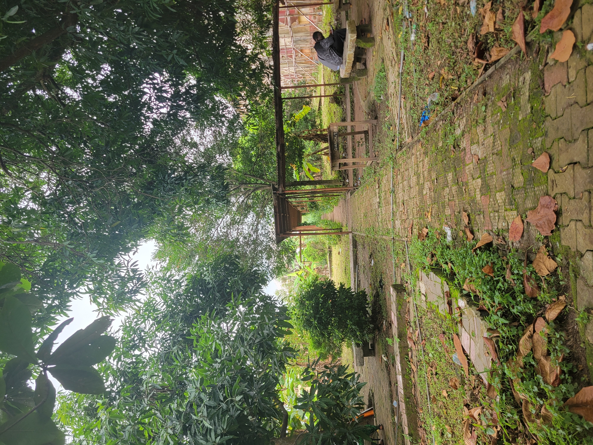 The image depicts an outdoor scene with a paved walkway leading through a leafy, green environment. The walkway is constructed of brick pavers with moss growing in the crevices. Flanking the walkway are patches of grass, fallen leaves, and scattered debris. On the right side, a person is seated on a stone bench, looking down at their hands, suggesting they are engaged with a device or object. Adjacent to the bench is a wooden table with some text that appears to be "D/ARCH/T/88" visible. A covered structure, possibly a shelter or canopy supported by poles, extends along the right side of the walkway. In the background, more trees and vegetation are visible, along with what appears to be a partially constructed building with scaffolding. The overall impression is one of a public outdoor space, perhaps a park or campus area, in Ukukwa, Nigeria.