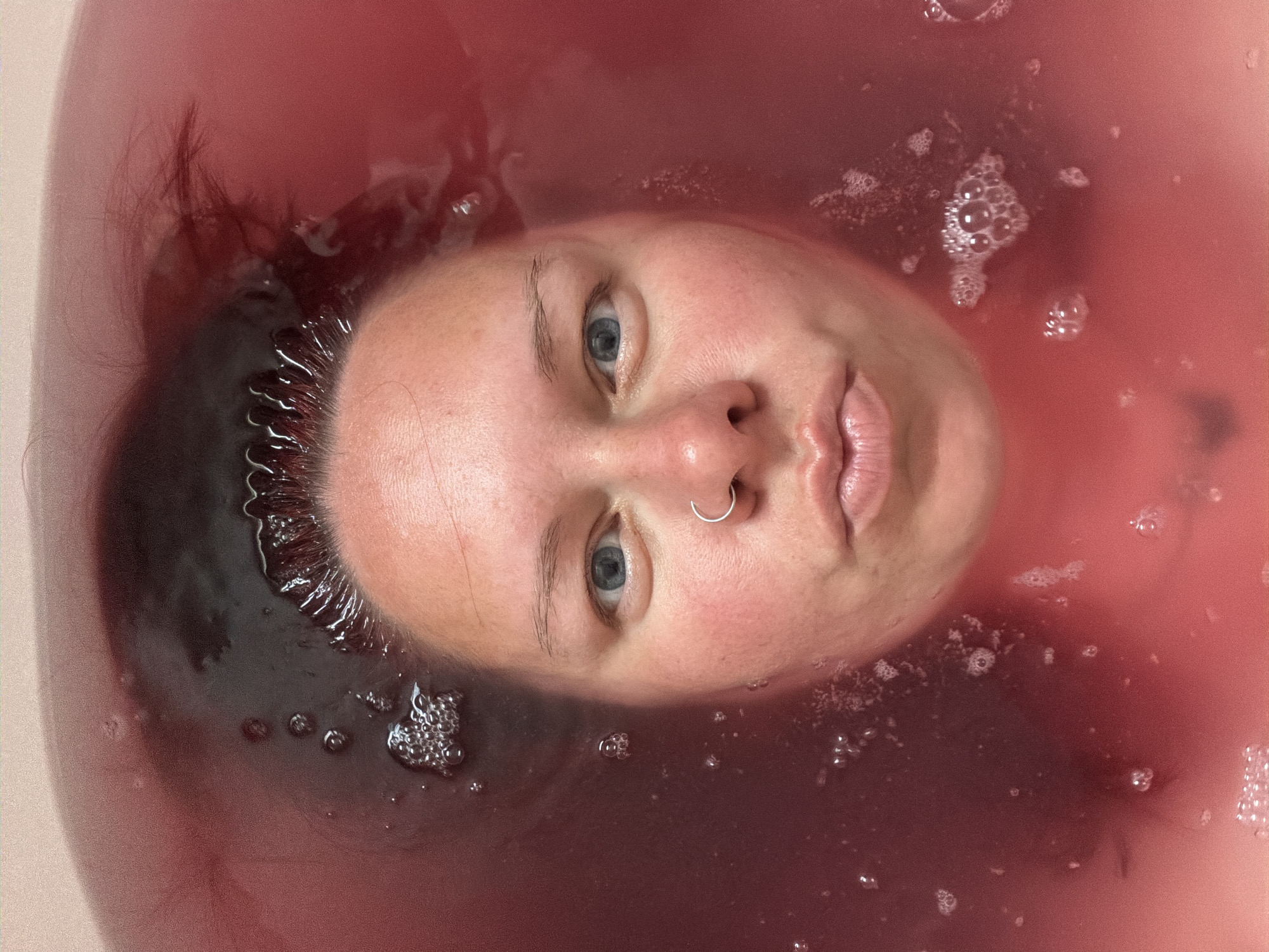 A person's face is floating in reddish-pink water. Bubbles are visible around the person's head, suggesting a bath or tub environment. The person has fair skin, light blue eyes, a silver nose ring, and dark hair partially submerged in the water. The location is not discernible in the provided image.