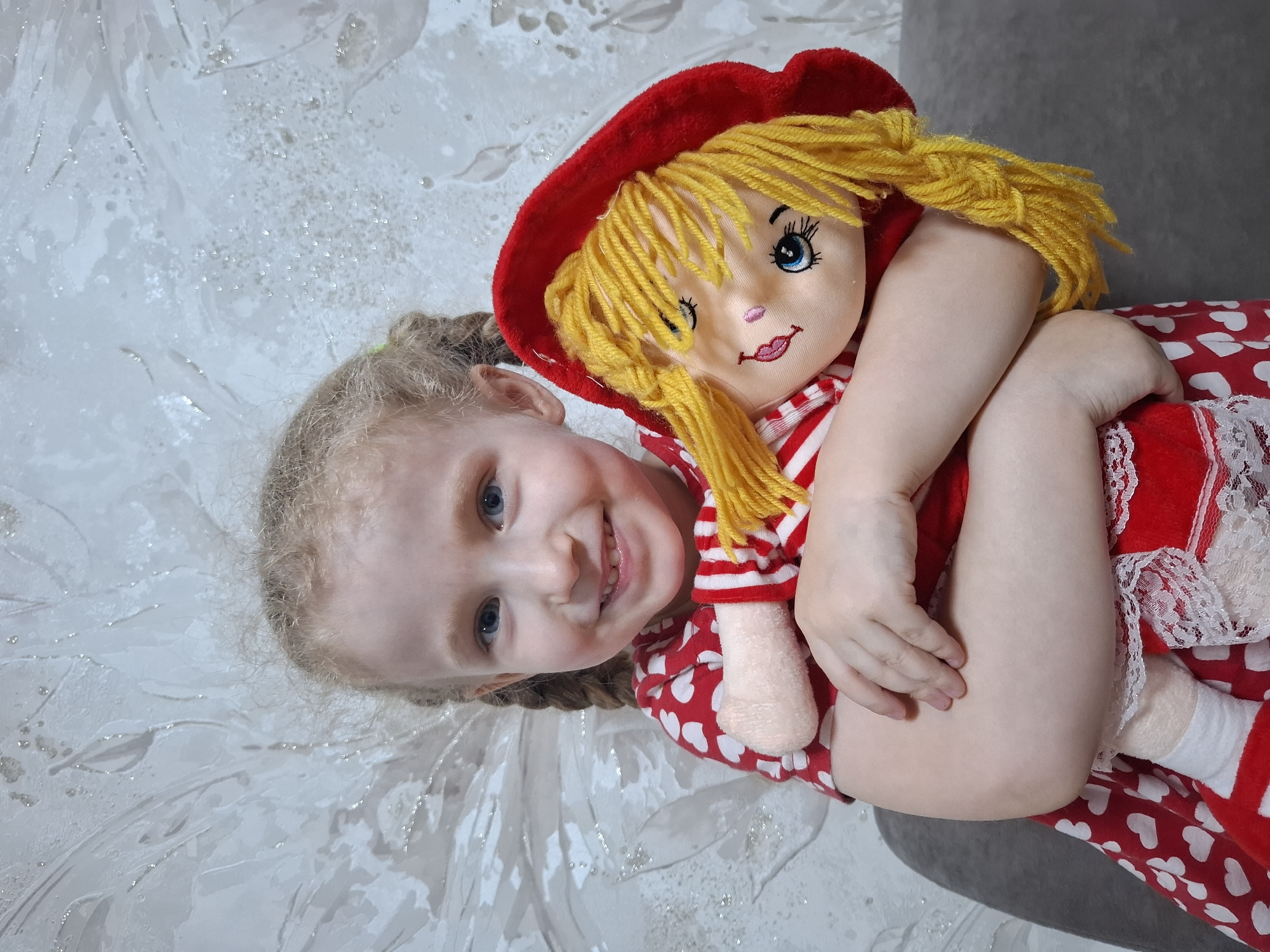 A young child with blonde hair, styled in pigtails, is lying down and hugging a doll. The doll has yellow yarn hair and a red hat. The child is smiling. Both are wearing red and white clothing. The background is a textured, light-colored surface. There is no evidence to confirm the location is Chaikovsky, Russia.