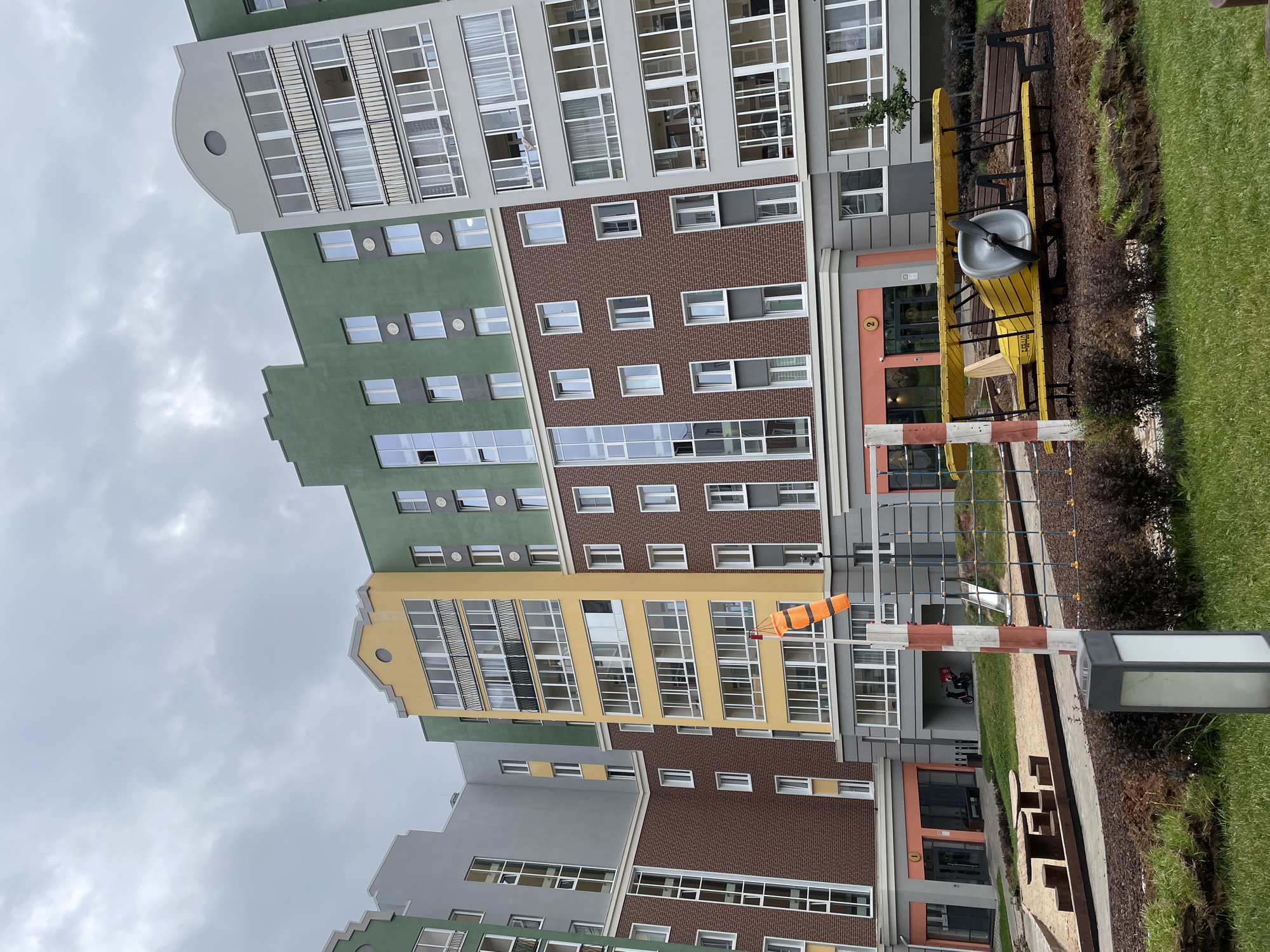 In the city of Pervomaysky, Russia, a modern residential complex is captured under an overcast sky. The complex features multi-story buildings with a varied color palette, including green, yellow, brown brick, and gray facades. Large windows and balconies are prominent architectural elements. In the foreground, a children's playground is visible with a sandbox, picnic tables, and a unique yellow biplane-themed play structure. A windsock, striped in orange and white, stands near a red and white striped pole. A small scooter with a delivery box is parked under an archway of one of the buildings. The grounds are landscaped with grass, bushes, and bare soil.