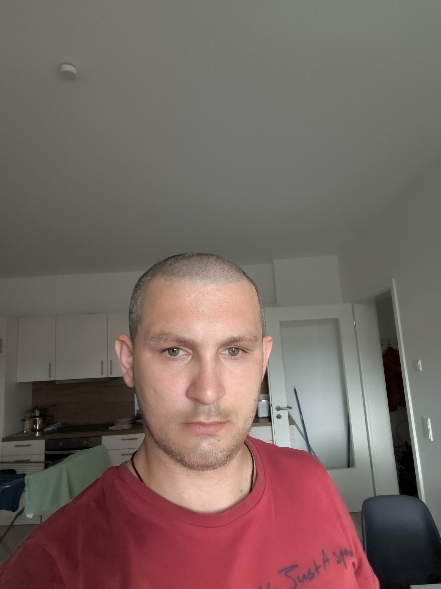 The image is a selfie taken indoors, likely in a residential setting in Wolgast, Germany. The subject is a fair-skinned man with a buzzed haircut, brown eyes, and stubble. He is wearing a red t-shirt with some writing on it. The background includes a kitchen with white cabinets and wooden countertops, a partially visible doorway, and a dark chair. A smoke detector is visible on the ceiling. The lighting suggests it is daytime.
