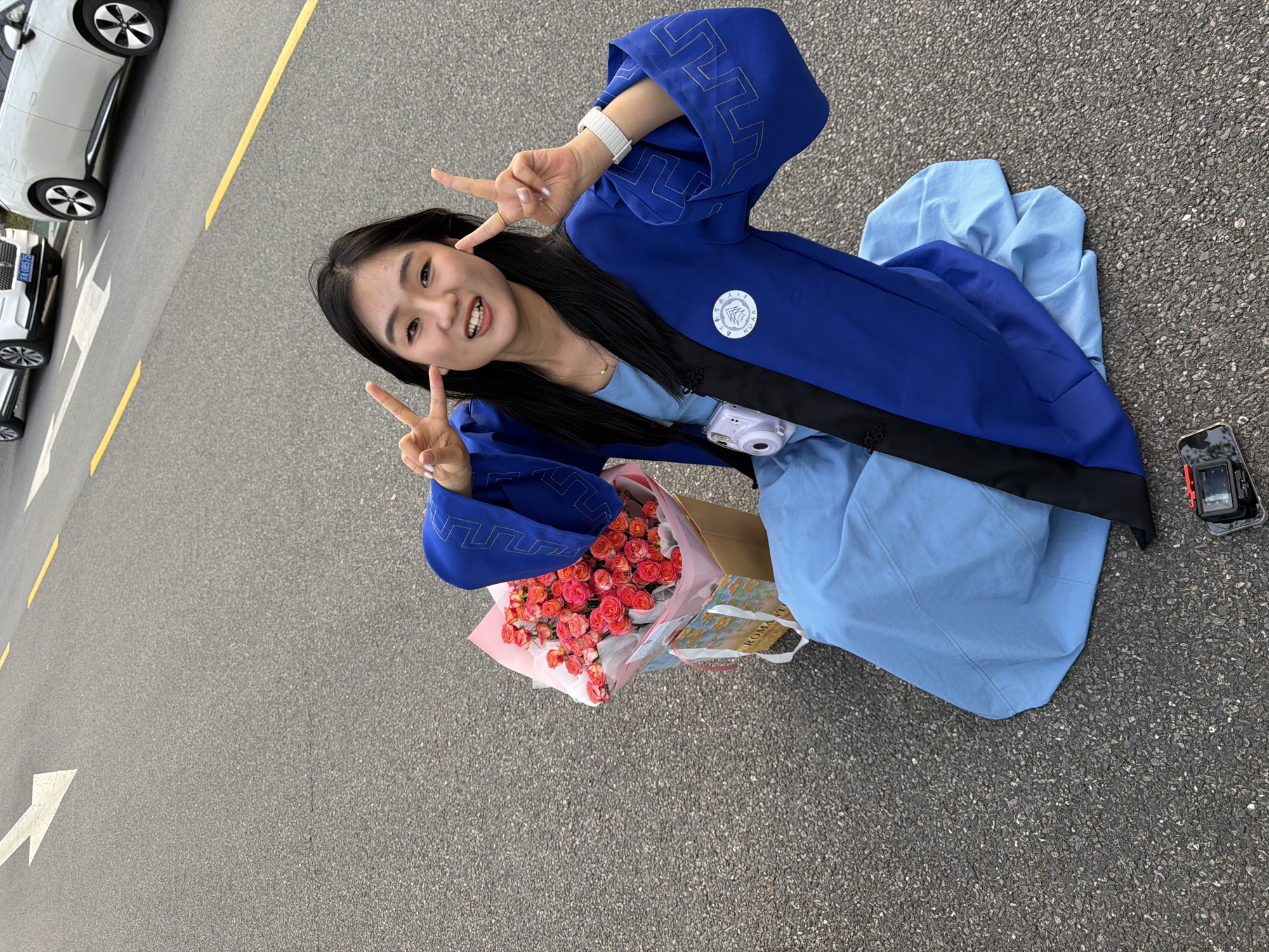 A young woman is sitting on the asphalt pavement in Jiangning District, China. She is wearing a light blue dress covered by a dark blue robe with silver geometric patterns on the sleeves and a round logo patch on the chest. A black band stretches diagonally across the robe. She is holding a bouquet of pink and red roses. Her black hair is long and she is smiling, making a peace sign with both hands. To her right is a black smartphone lying face up on the pavement. In the background, there are parked white cars and a yellow lane divider. A white arrow is painted on the pavement.