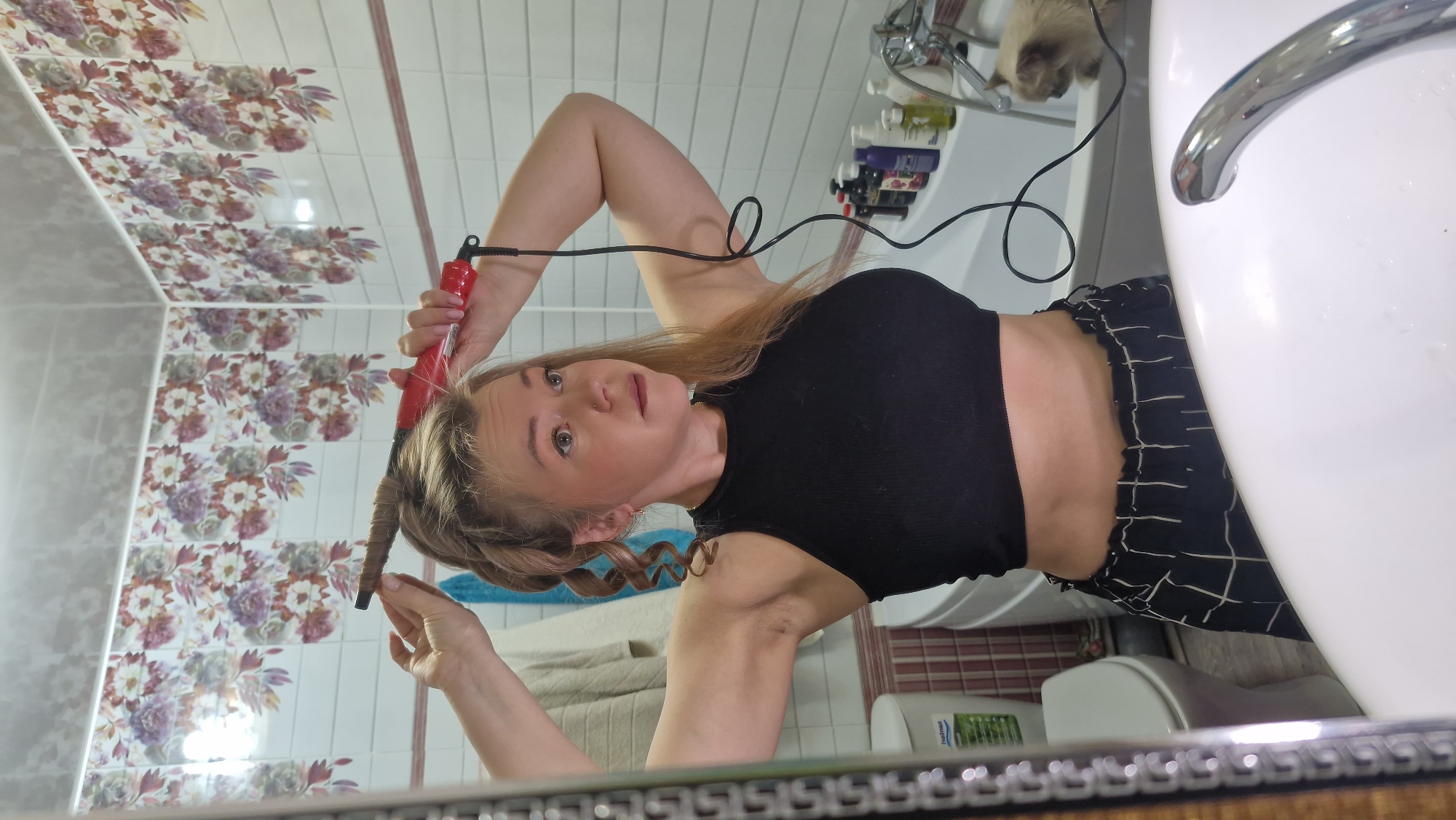 A selfie captured in a bathroom mirror in Otchor, Russia, shows a young woman in the process of curling her hair. The image is oriented vertically, with the woman looking directly into the mirror, holding a red-handled curling iron to a section of her blonde hair, which is wrapped around the barrel to form a curl. She wears a black ribbed crop top and black pants with a white grid pattern.
The bathroom setting is evident from the white sink and chrome faucet visible on the right, and various bottles (shampoo, conditioner, etc.) lined up on a shelf or counter behind her. A light-colored cat with tan fur can be seen curiously peeking from behind the sink in the upper right background. The bathroom walls feature distinct floral-patterned tiles in shades of purple, pink, and green, interspersed with plain light grey tiles. A light-colored towel is draped in the background, and a white toilet or bidet is partially visible in the reflection. The floor, also seen in reflection, appears to have a reddish-brown striped tile pattern. The woman has a neutral to slightly focused expression. Visible text on a bottle in the background reads "homise".
