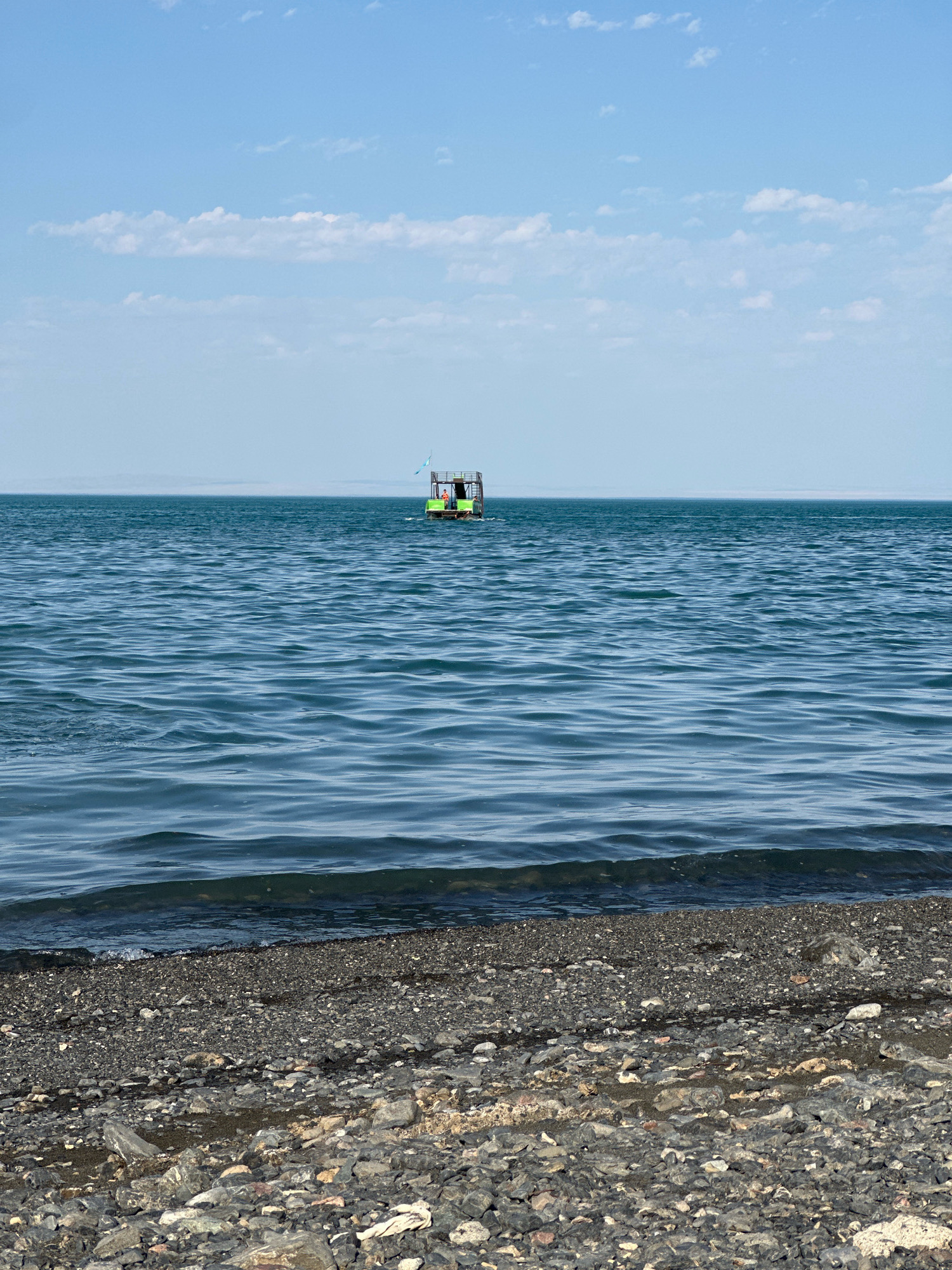 The image depicts a serene body of water, likely a lake, with gentle ripples on its surface. In the distance, a small, green structure with a light-colored roof is floating on the water. This structure appears to be a boat or raft, possibly used as a shelter or a recreational spot. Two small flags, one blue and white, are attached to the structure. The sky above is a clear, pale blue, with a few scattered white clouds. The foreground is a shoreline covered with dark, grey pebbles. The overall scene suggests a tranquil outdoor setting, possibly a lakeside resort or a natural landscape in Көктұма, Kazakhstan.