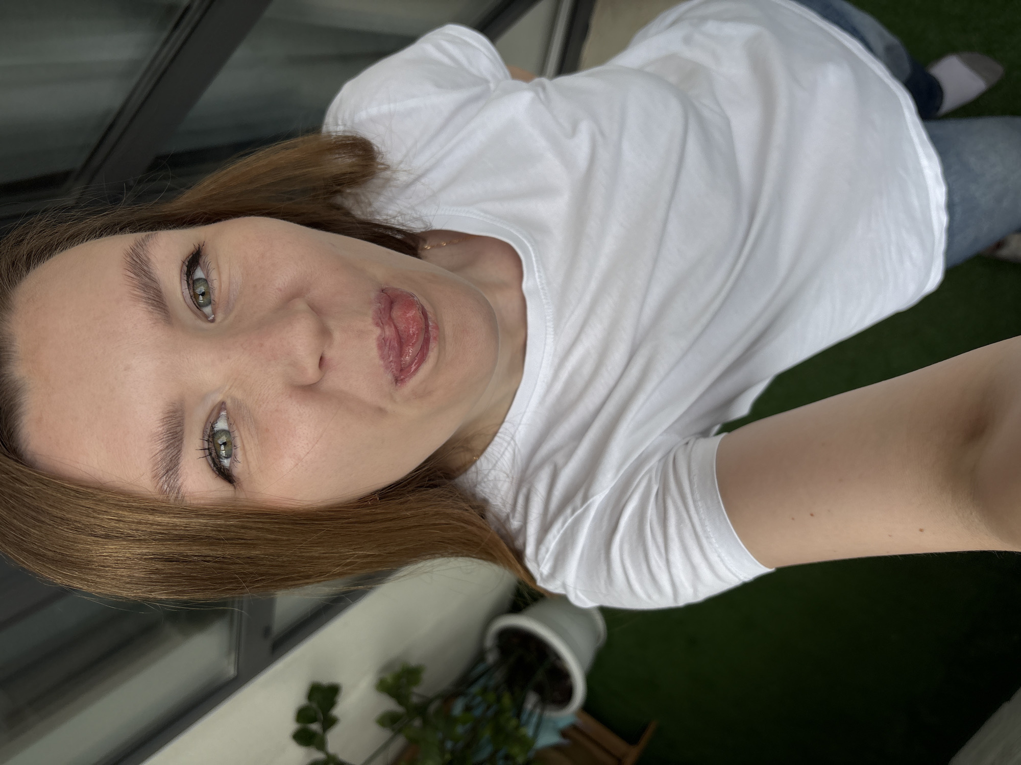 A woman with shoulder-length auburn hair and green eyes is taking a selfie outdoors. She is wearing a white t-shirt and blue jeans. Her tongue is sticking out, and she has a playful expression. In the background, there is a potted plant and a railing, suggesting she is on a balcony. The setting appears to be a residential area.
