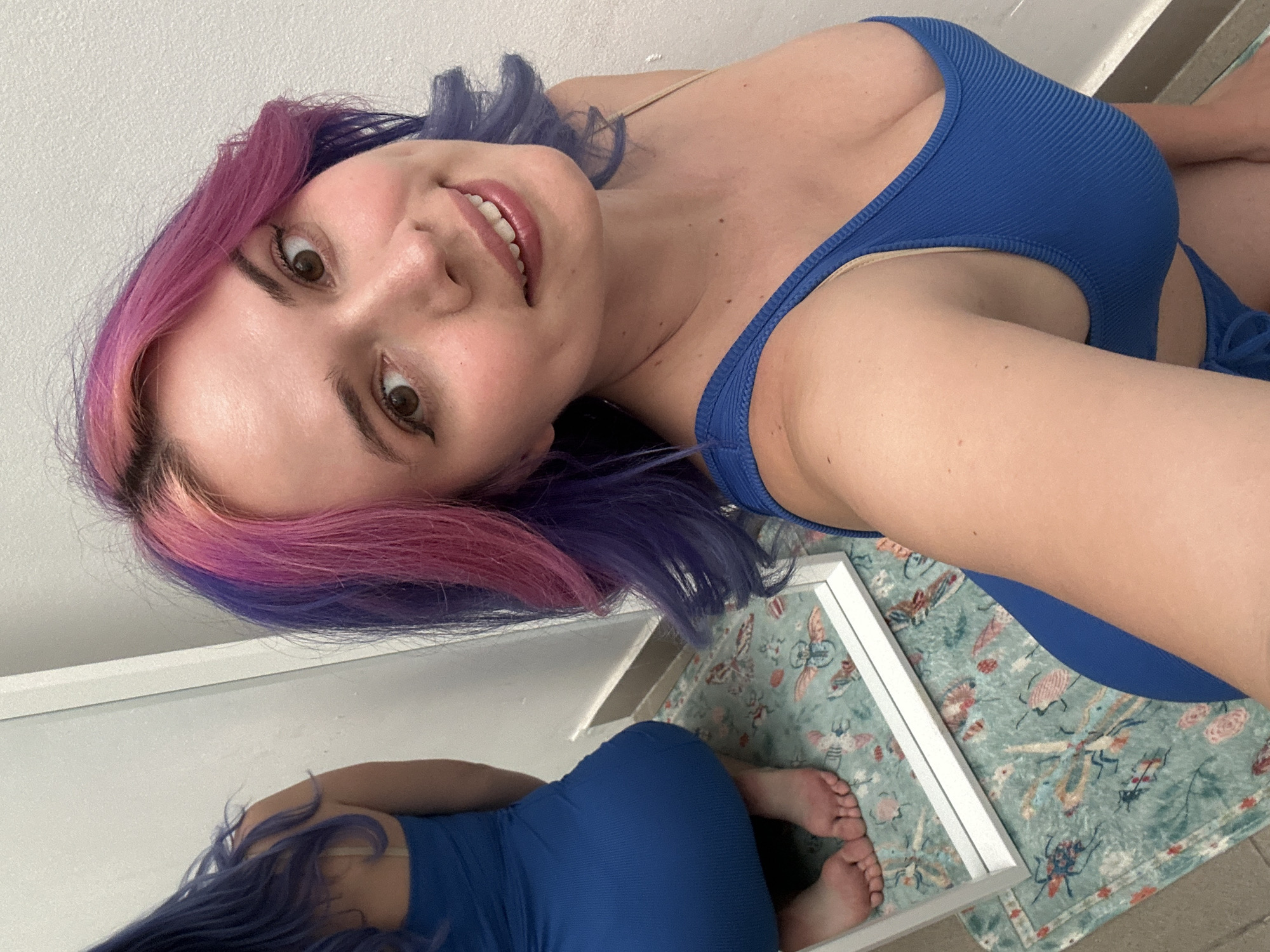 This media file captures an indoor selfie of a young woman, taken at an angle that positions her head towards the top right and her body extending downwards. The woman has distinctive two-toned hair, featuring vibrant pink on the front sections and purple further back. She is smiling broadly, revealing white teeth, and looks directly towards the camera. Her eyes are brown, and she has visible eyeliner.
She is wearing a blue, ribbed garment, appearing to be a swimsuit top or a form-fitting tank top, which covers her torso. Her skin is fair.
A large rectangular mirror is visible in the lower part of the frame, angled towards the floor. The mirror reflects the lower portion of her body, showing her hips, legs, and bare feet, all encased in the same blue, ribbed material. Also visible in the mirror's reflection is a light green mat or rug adorned with a repeating pattern of various insects and butterflies in different colors. The background behind the woman is a plain white wall.
The image conveys a cheerful and casual mood. There is no visible text. The location context provided is the City of Zagreb, Croatia, though no specific geographical markers are visible in the indoor setting of the image itself.