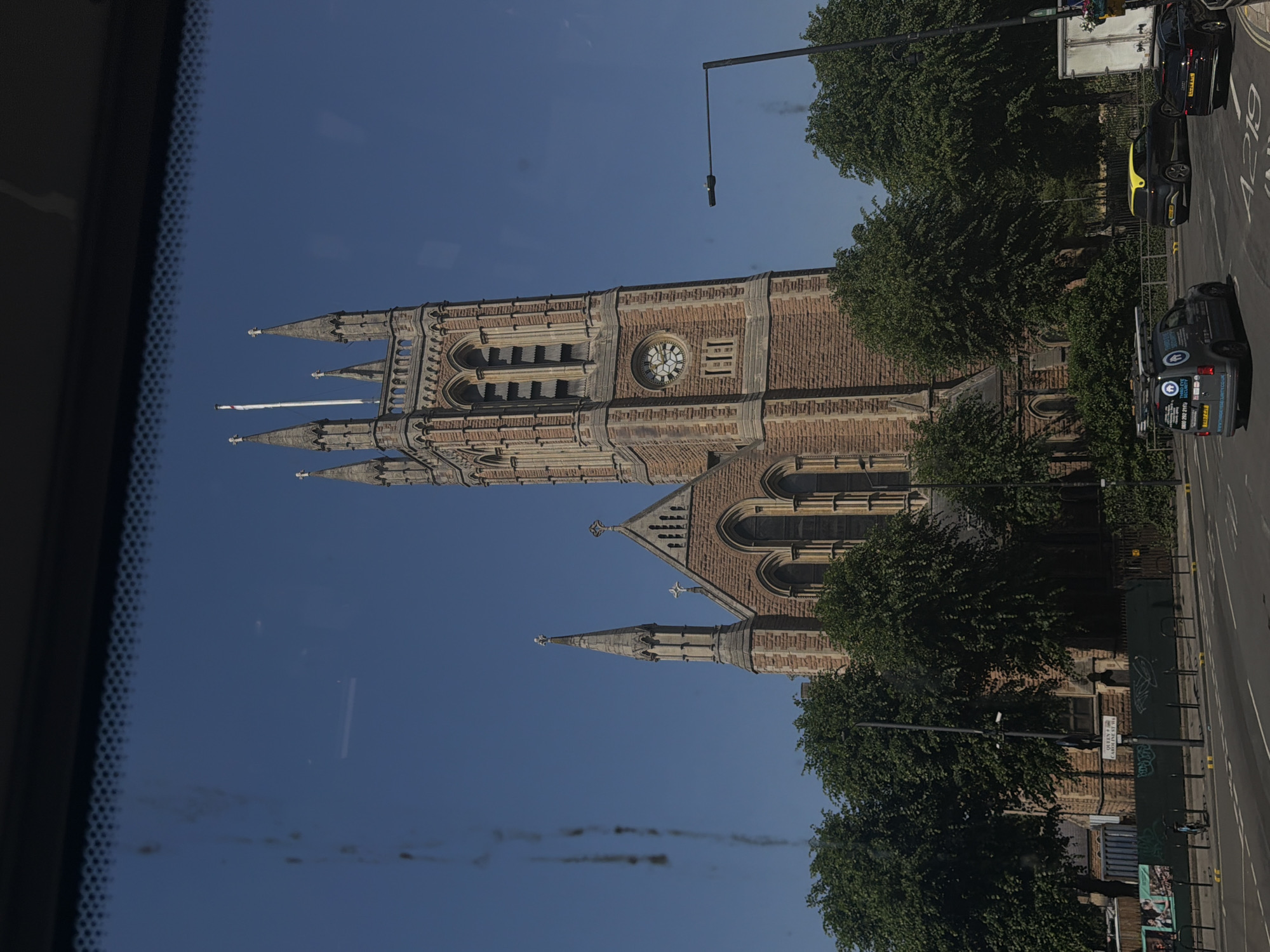 The image shows a view of St Mary Redcliffe Church in Bristol, United Kingdom. The church is a large, gothic-style building with a tall spire, visible on the left side of the image. The church is constructed from dark red stone with gray stone accents. A clock is visible on the tower. A row of cars is visible on the right side of the street along with the street sign "West Caroline Place". The image appears to be taken from inside a vehicle, with the edge of the windshield visible on the left side of the frame. The sky is a clear blue. Trees line the street on the right side of the frame.
